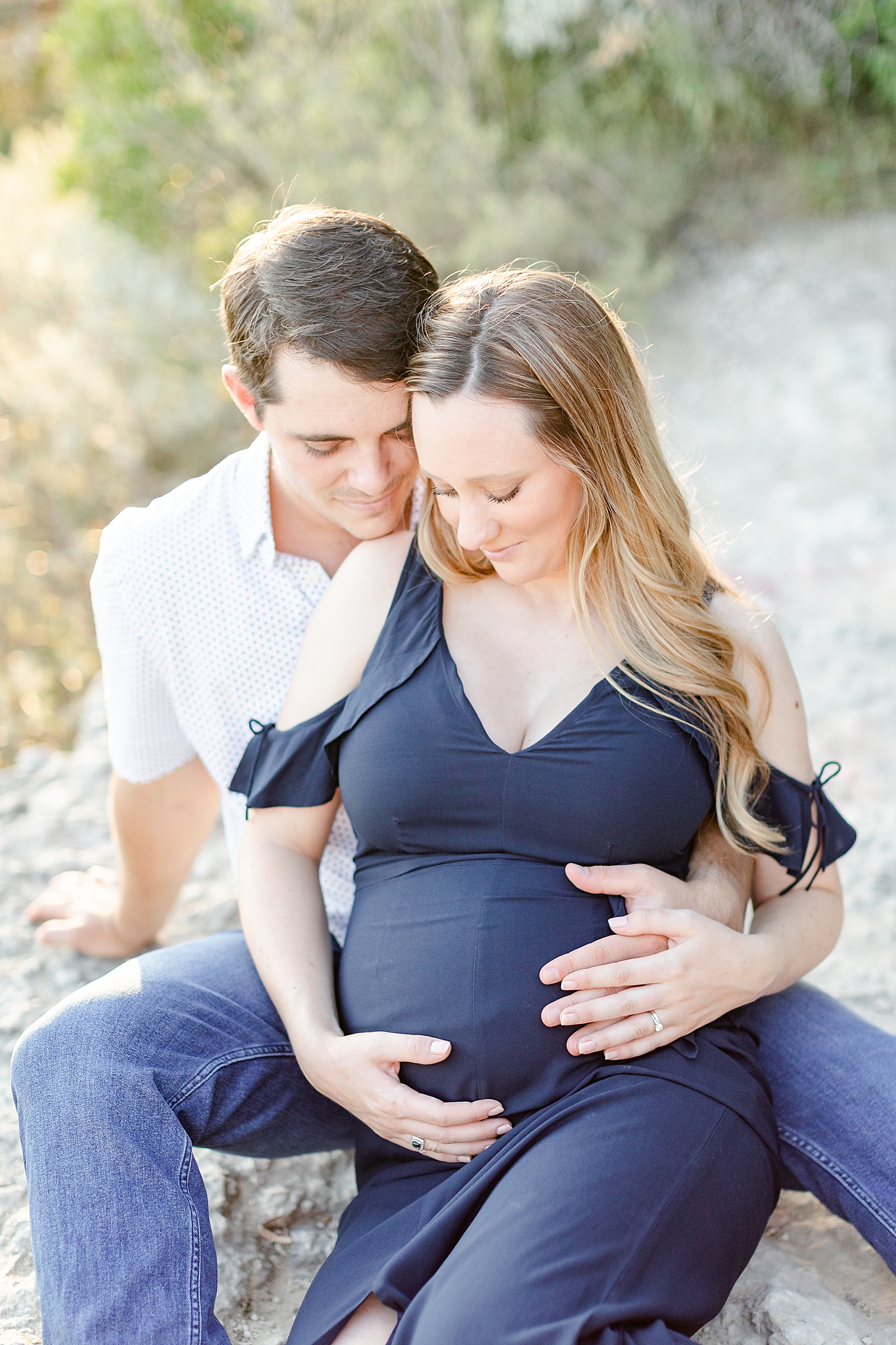 Pregnant mom in a navy blue maxi dress sitting between husbands legs holding her baby bump while they both look down at belly