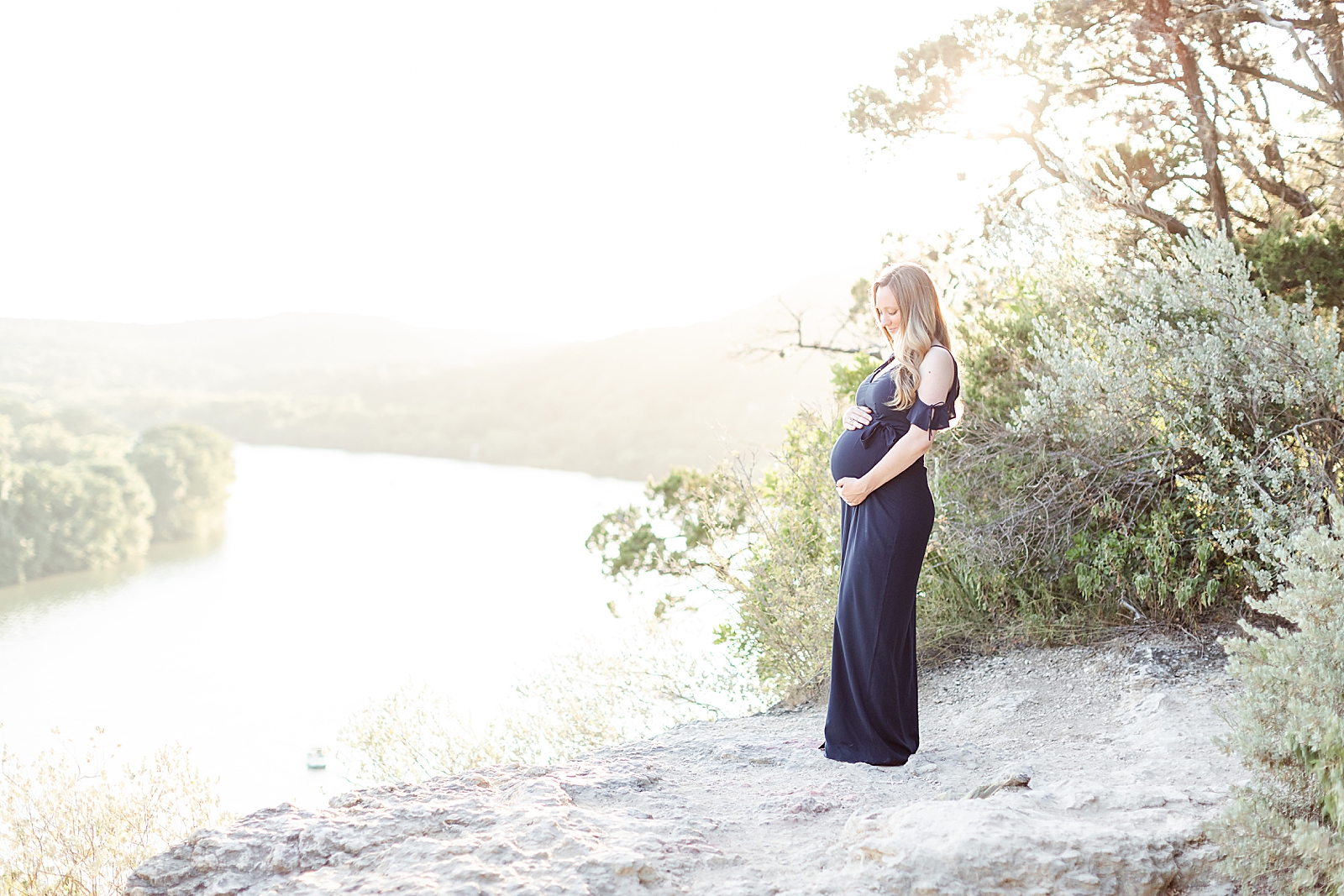 blonde mom wearing a navy blue maxi dress holding on to her baby bump looking down with lake Austin in the background at sunset