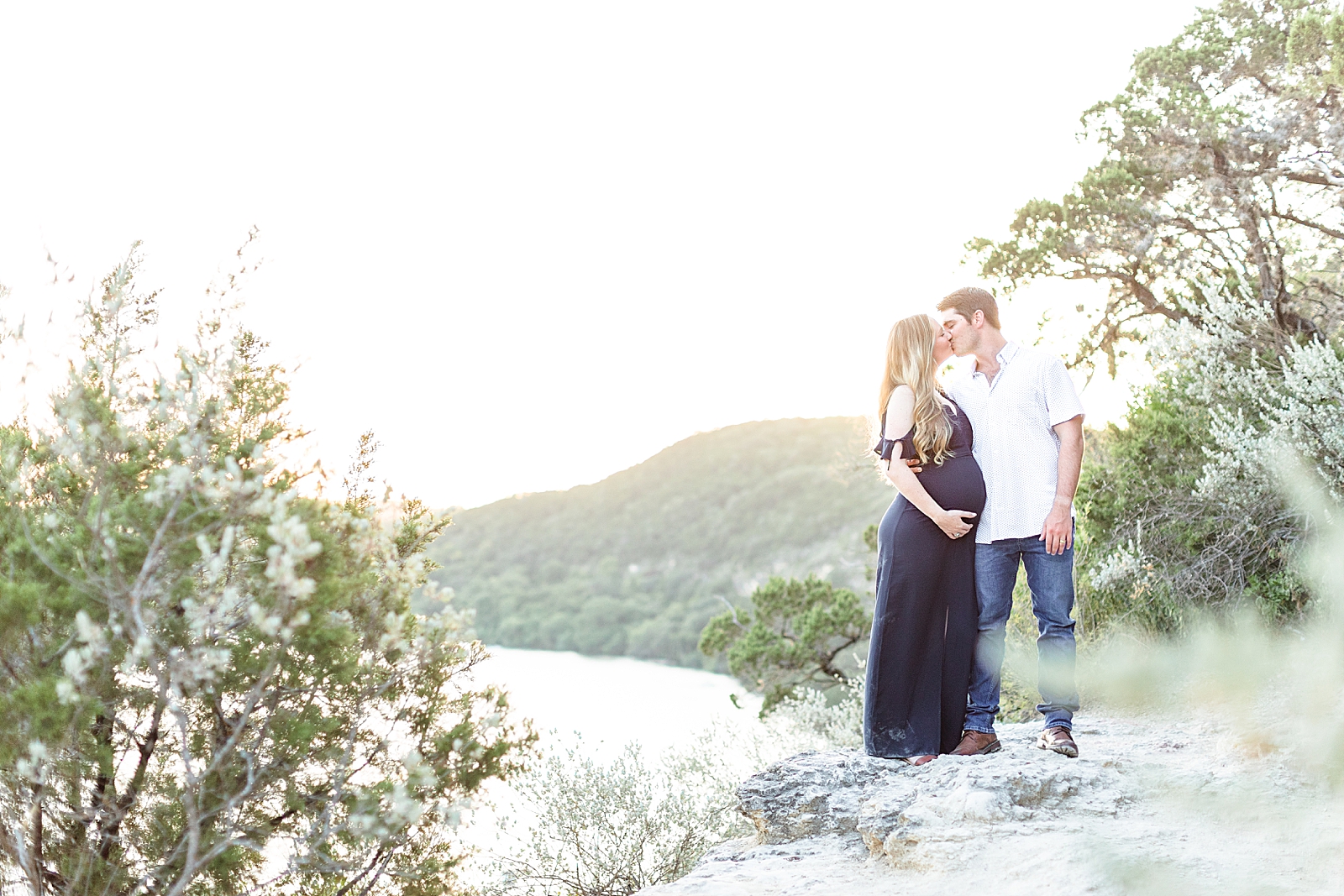Mom and dad to be kissing overlooking lake Austin at sunset with mom wearing a blue maxi and dad in a white button down and jeans