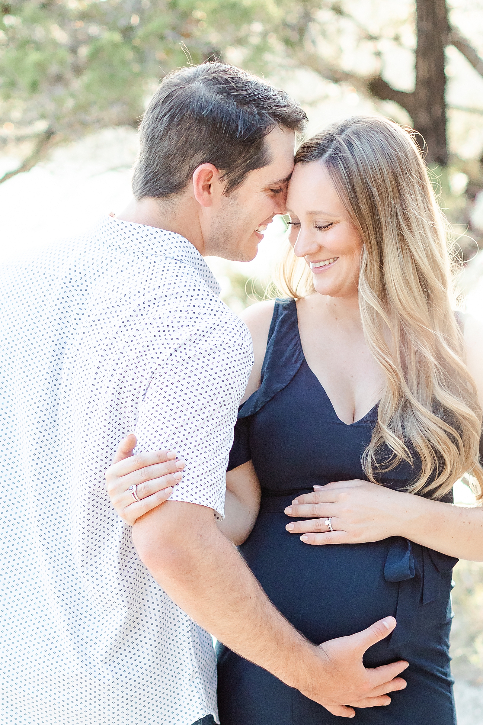 picture of mom and dad to be mom facing camera wearing navy blue maxi dress while husband faces away from camera with nose to his wife's temple reaching down and holding the baby bump