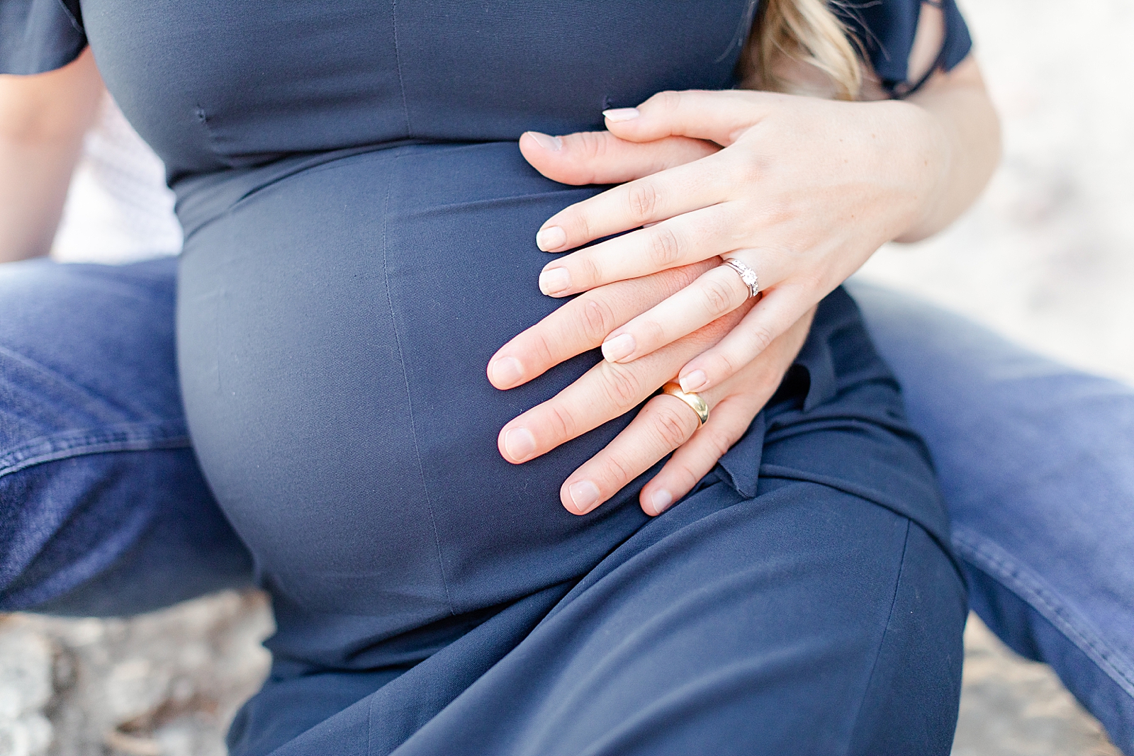 close up of husband and wife hands with wedding ring holding on to baby bump wearing navy blue dress