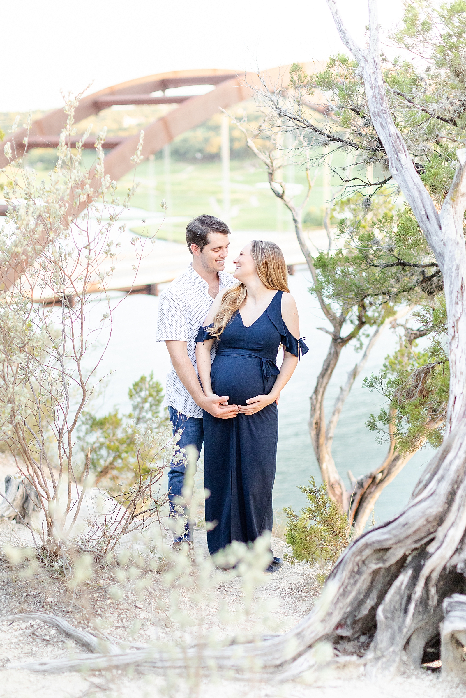 Husband and wife standing in front of the 360 bridge in Austin holding baby bump framed by a tree smiling at one another