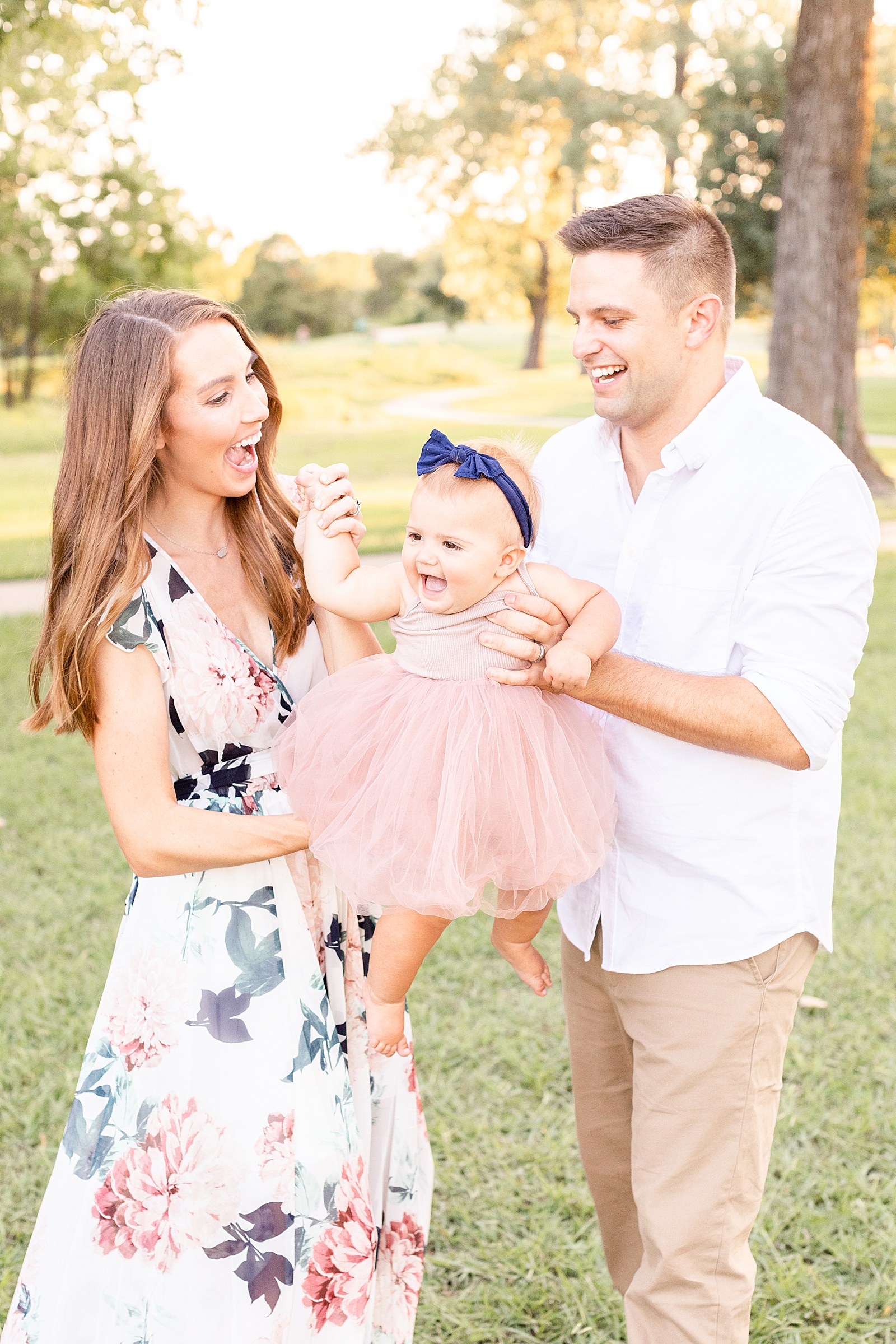 mom and dad hold baby and swing her around to get her to smile wearing her pink tutu dress and navy blue bow
