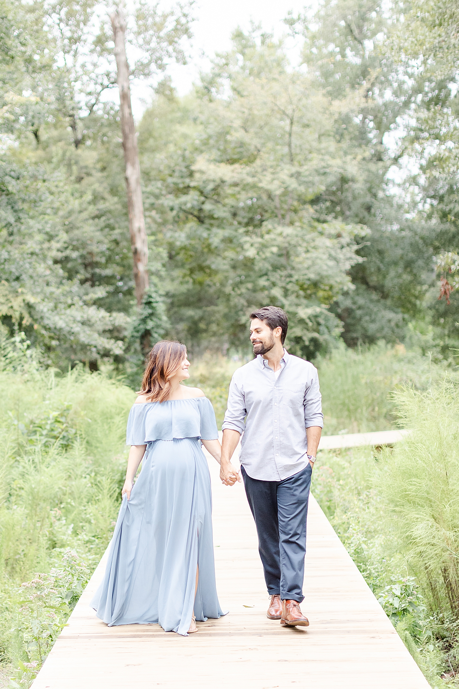 pregnant mom and husband walking down wooded pathway holding hands and smiling at each other while mom wears a blue gown at maternity session