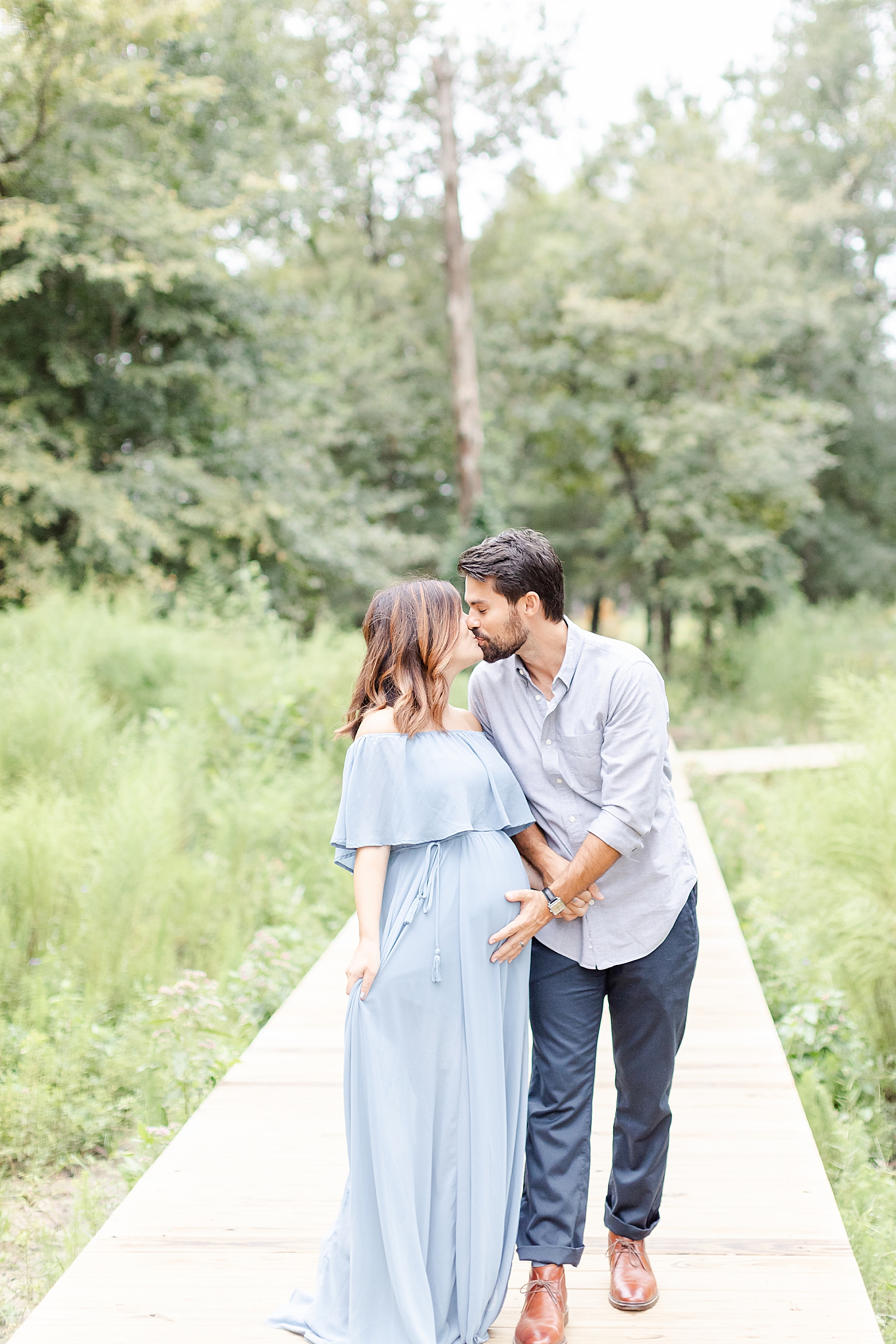 couple kissing at the end of a wood pathway surrounded by trees during rainbow baby maternity session