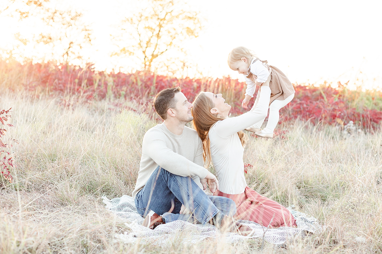 Pulled back family photo of mom and dad sitting on blanket and mom holding toddler daughter up in the air while she laughs mom and dad both looking at her smiling during their fall family session