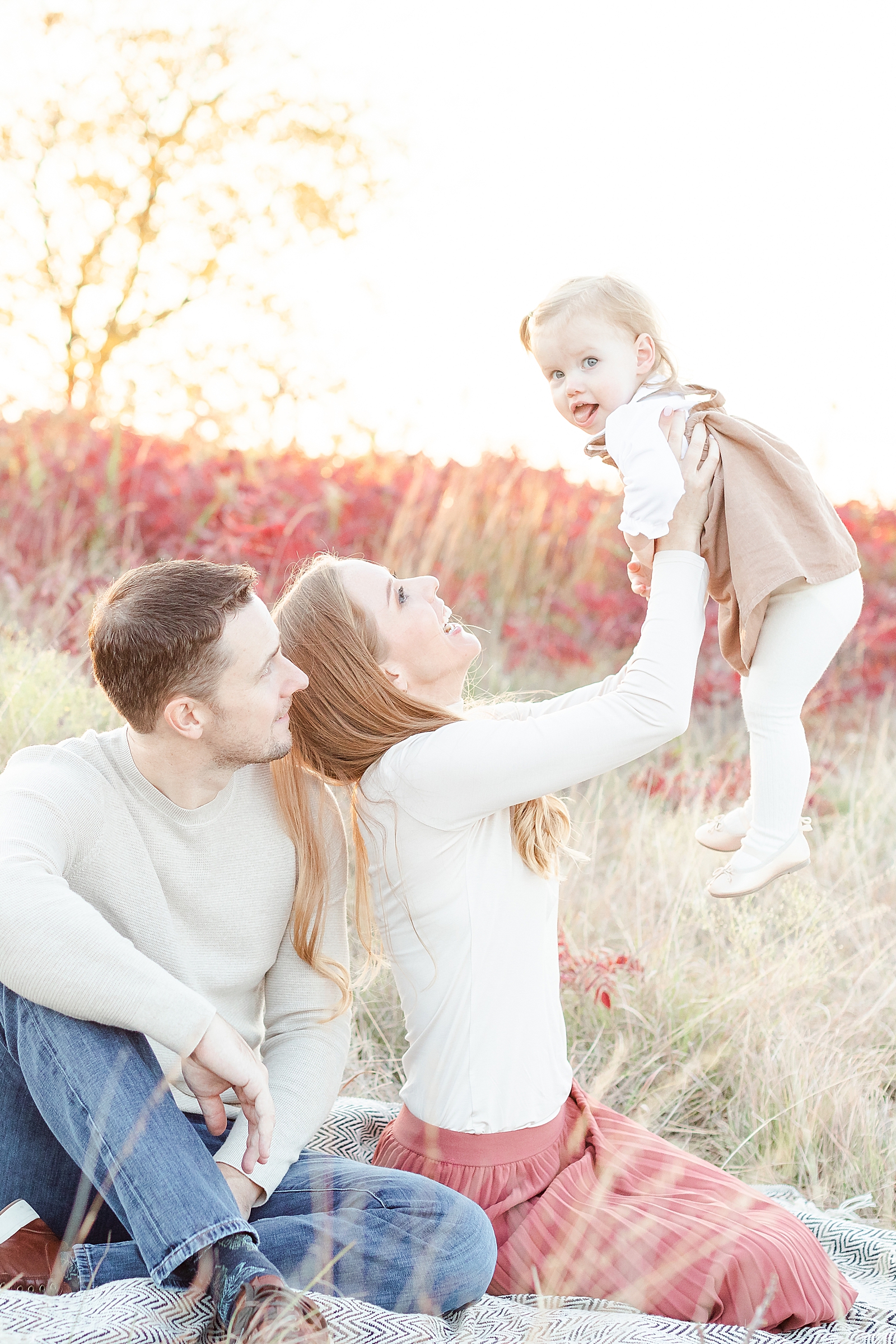 fall family photos with mom and dad sitting on blanket in a field during sunset holding toddler daughter up who is making a silly face while they smile at their daughter
