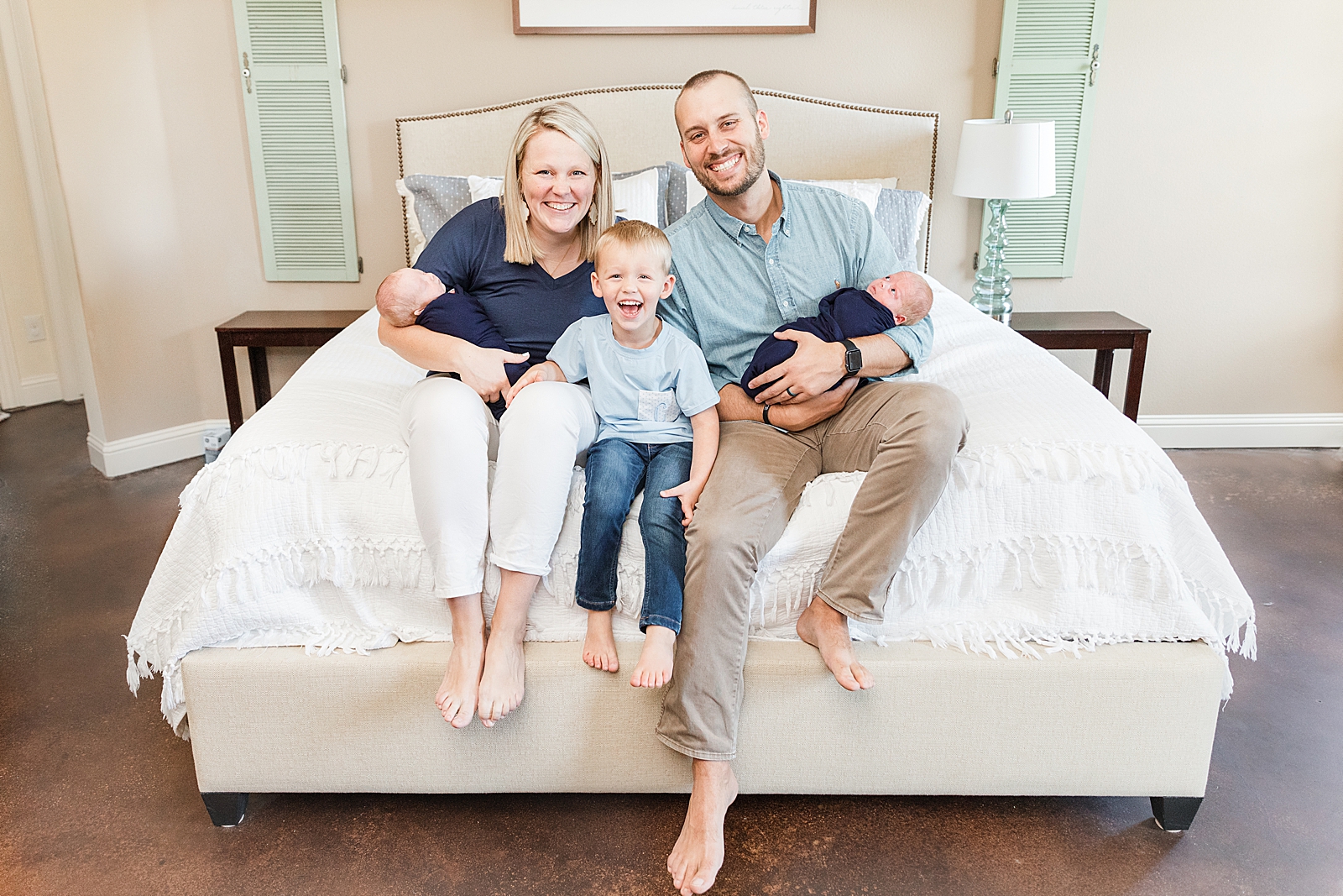 mom and dad and toddler boy sitting at the end of the bed smiling at the camera during their lifestyle newborn photos