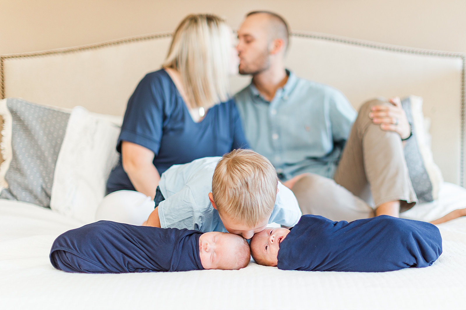 mom and dad share a kiss out of focus in the background while toddler boy kisses his twin newborn brothers on the head in front of them during lifestyle photos