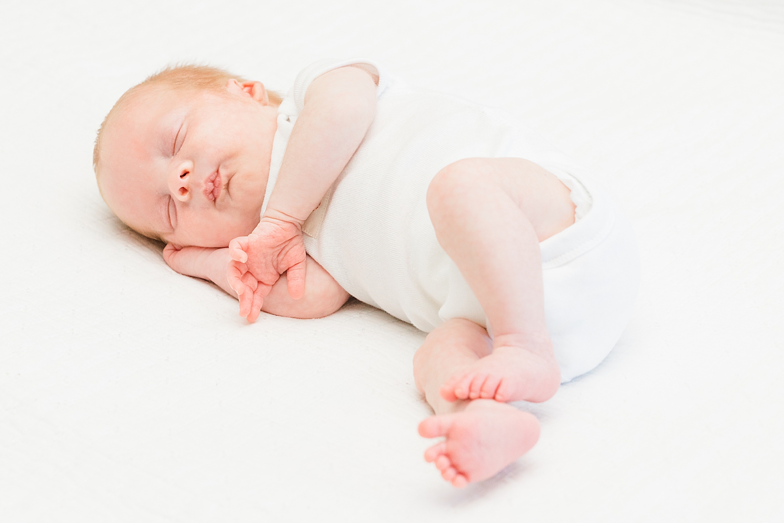 newborn baby in white onesie asleep on the bed during lifestyle newborn photos