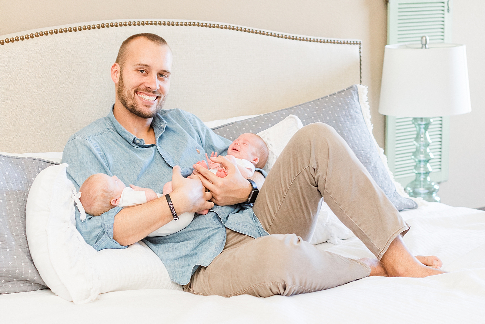 Dad laying back on bed in a blue button up shirt holding twin boys smiling at the camera during newborn photos