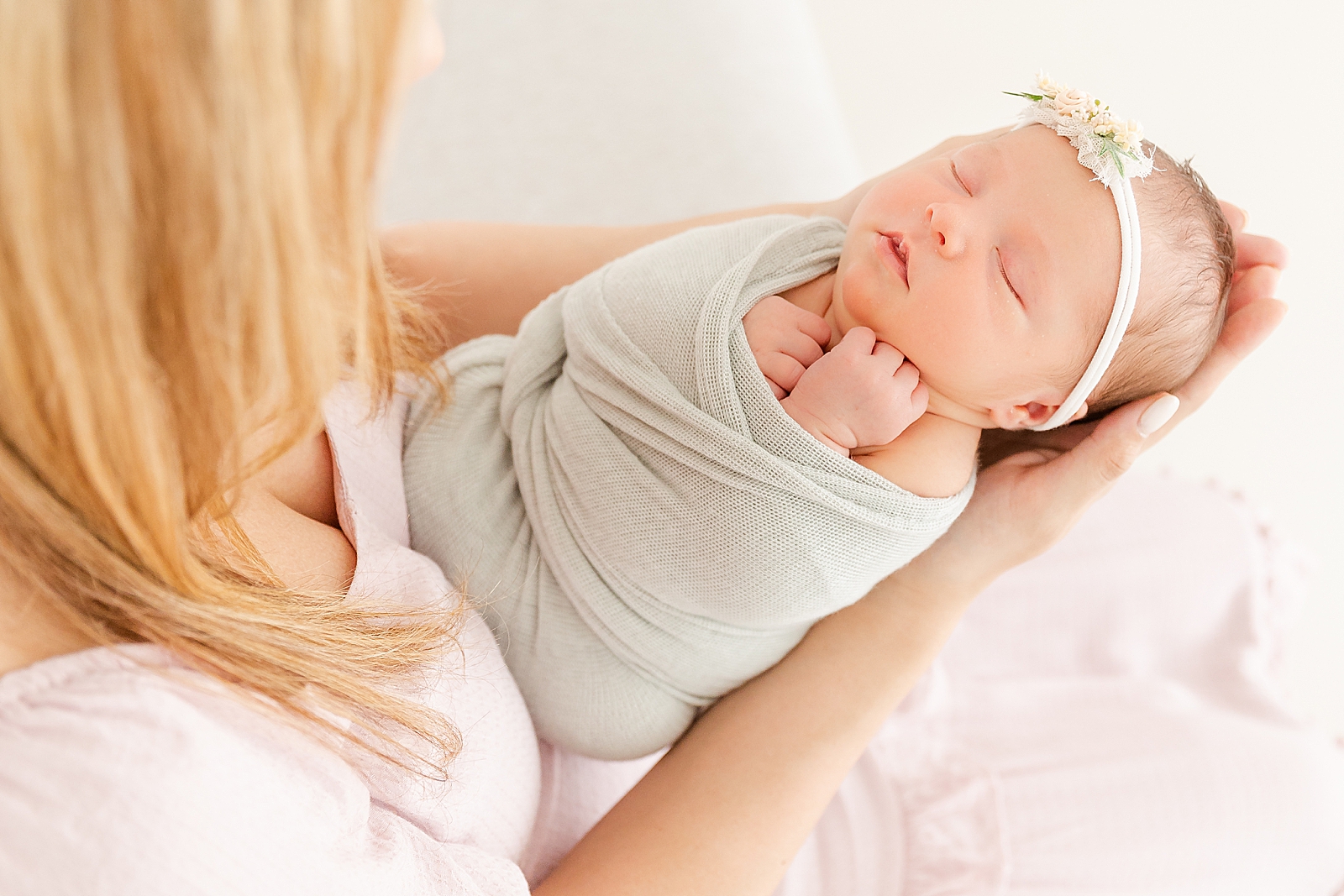 Mom holding swaddled newborn baby out in front of her while she gazes down at her wearing a sage green swaddle and flower headband