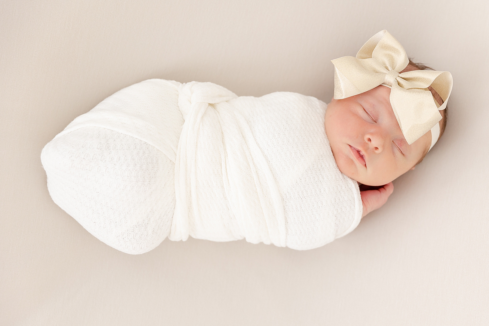 swaddle posed newborn image of baby wearing white swaddle with gold bow on camel colored background asleep