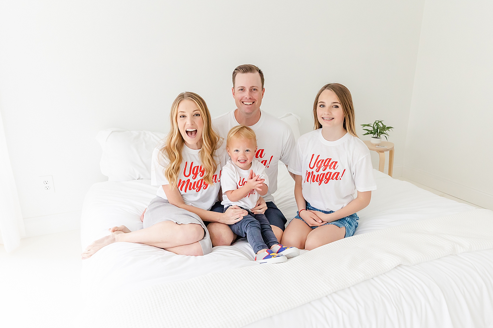 Family photo of a family of 4 sitting on a bed smiling at the camera wearing matching Daniel tiger ugga mega shirts during second birthday infertility photoshoot