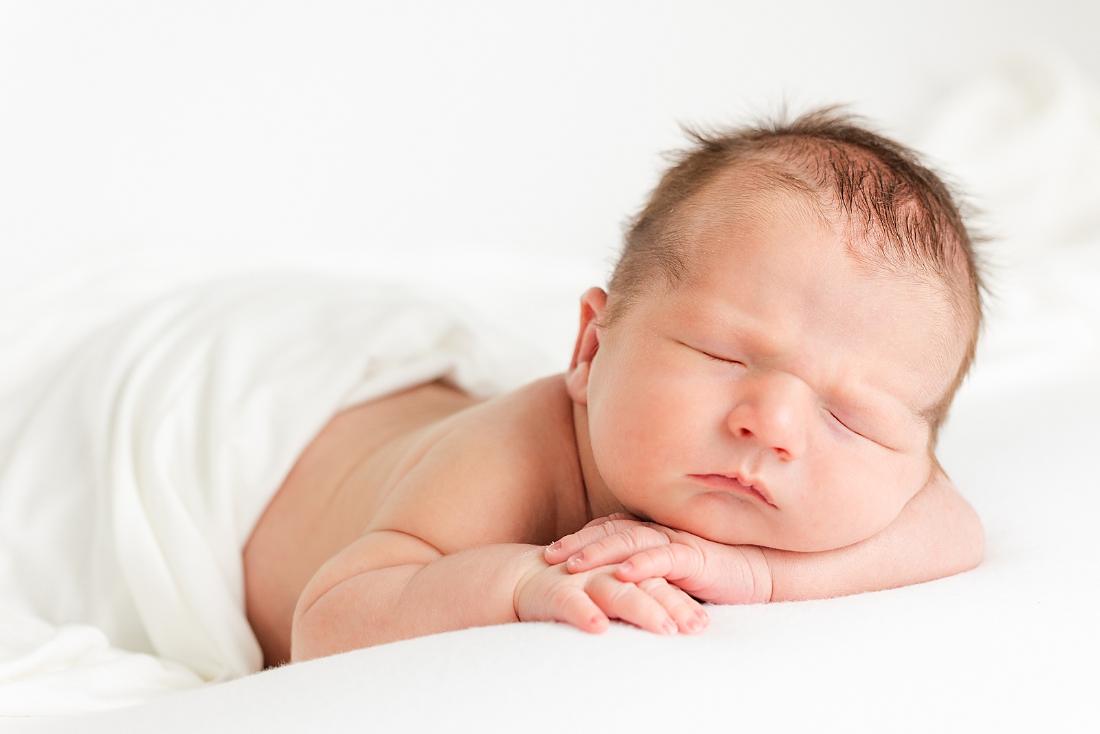 Posed newborn photo of baby sleeping with hands resting under shin on white background and white blanket draped over him