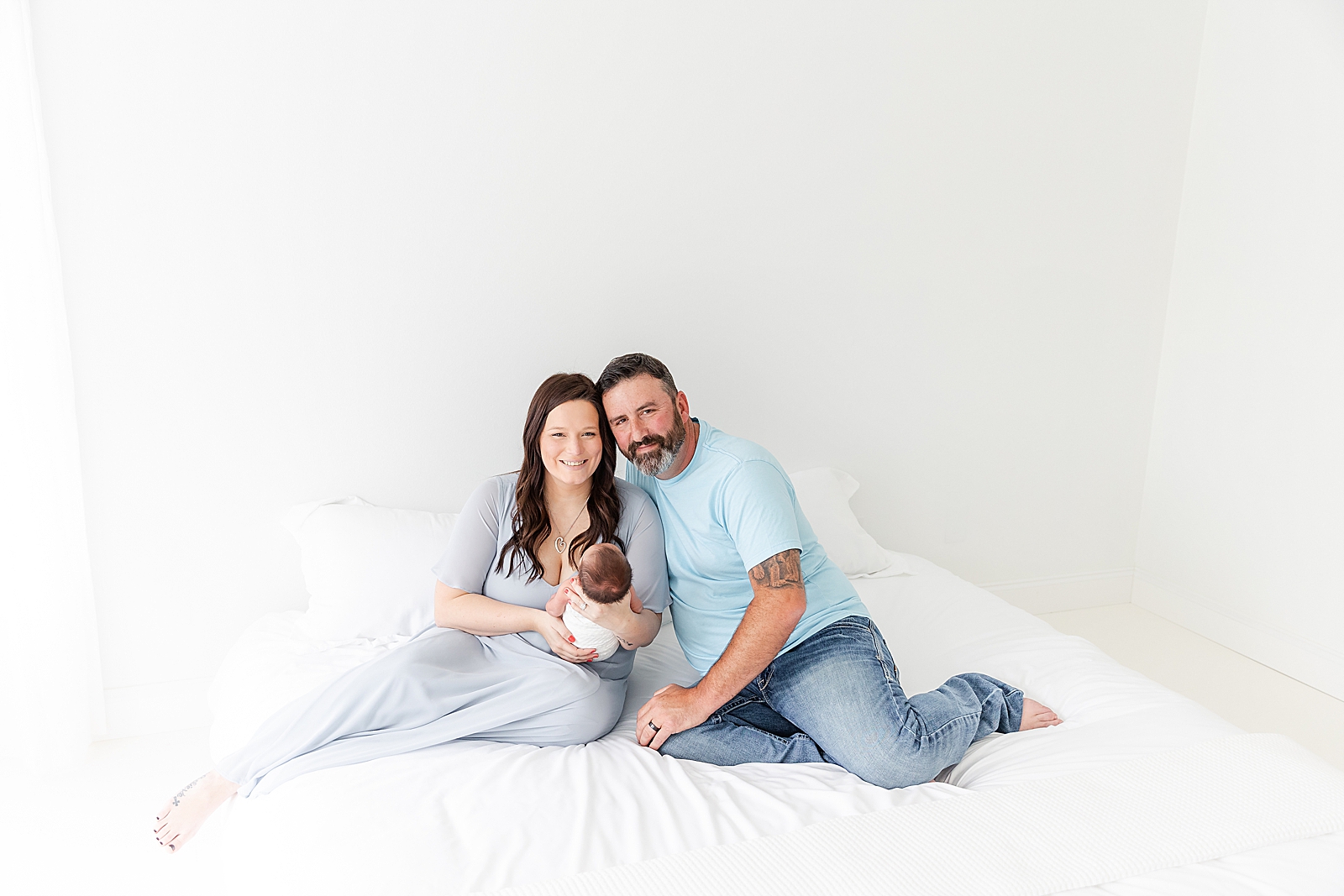 mom and dad sitting on white bed in studio holding baby smiling at camera for newborn photos