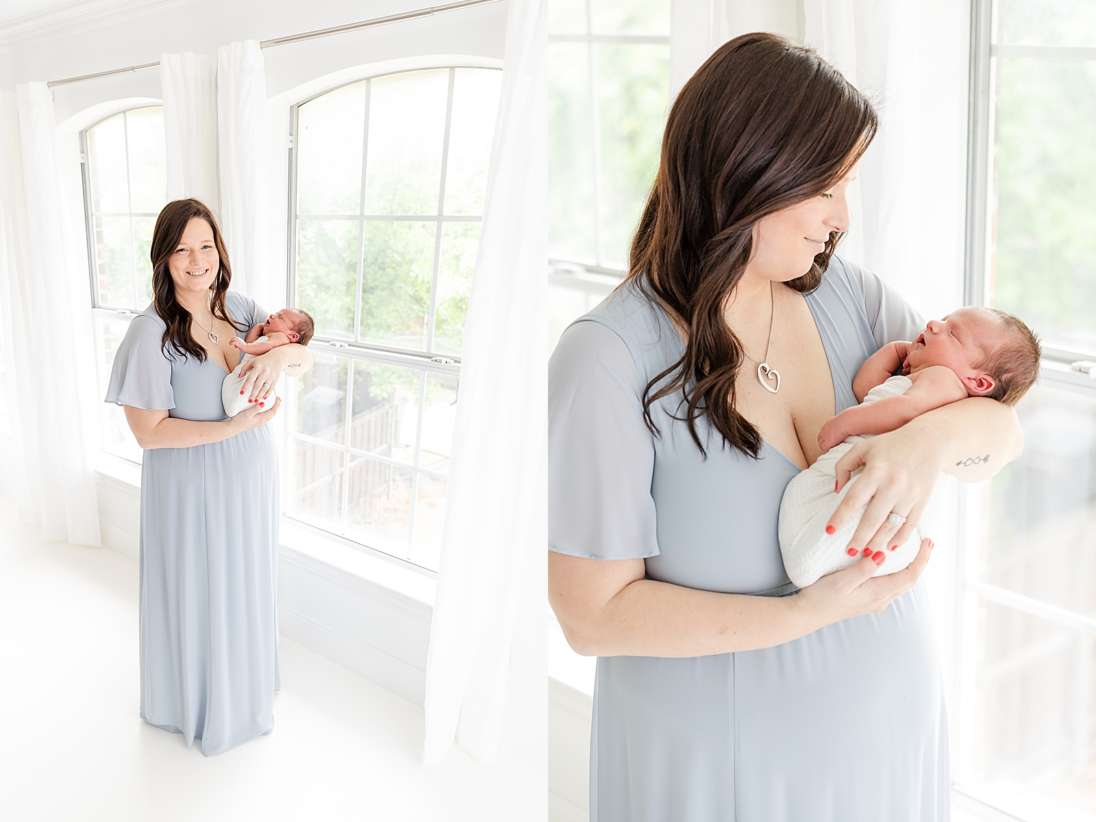 mom wearing a light blue gown for her newborn photos standing by window holding baby smiling at camera and smiling down at her newborn