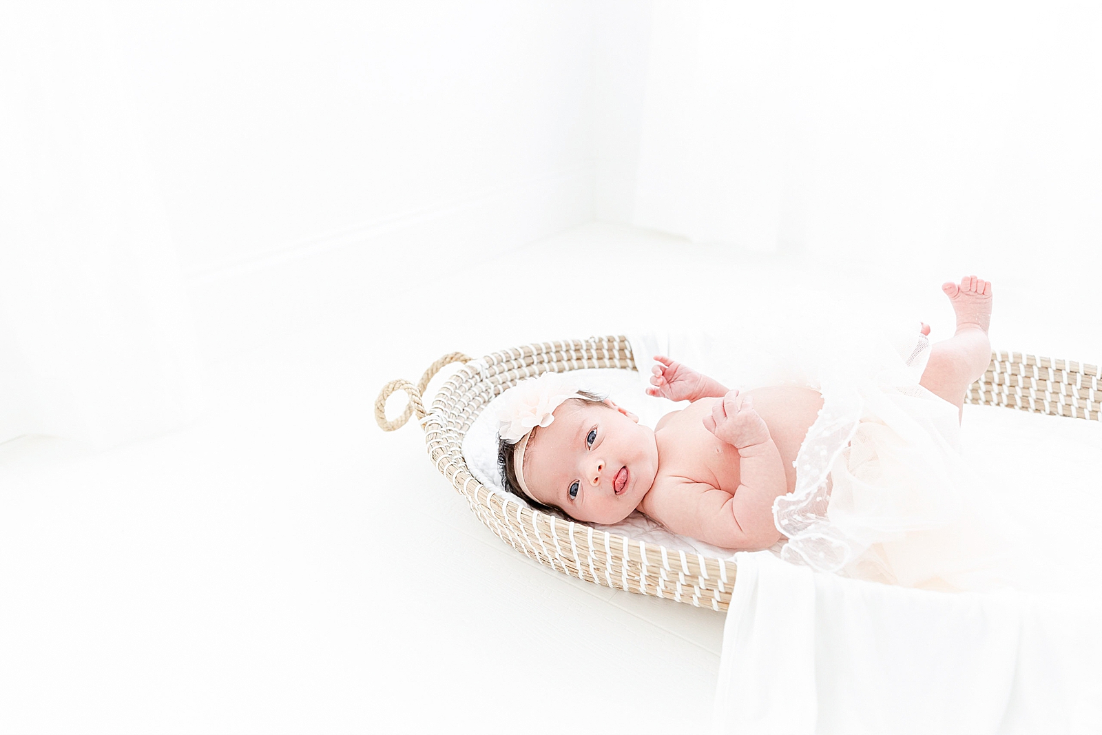 Newborn in Moses basket looking at the camera wearing a tutu and bow sticking her tongue out with feet up in the air