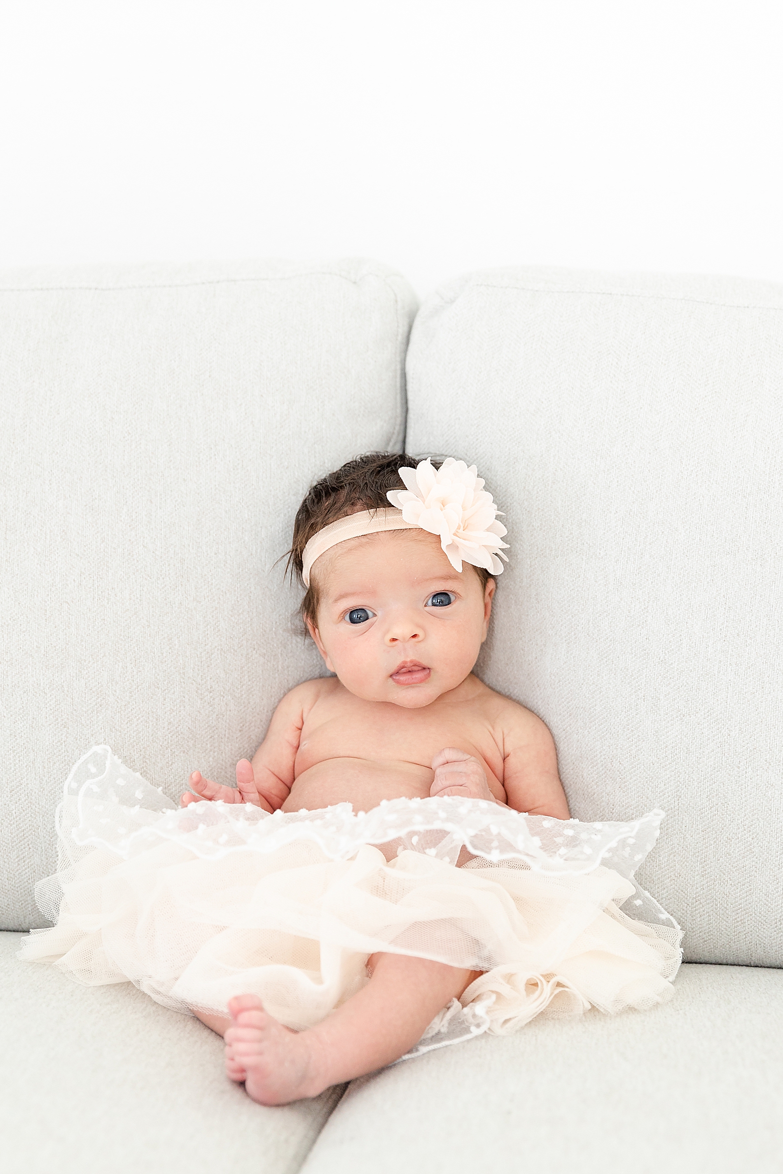 Newborn Sitting up on grey couch in a tutu and pink flower bow looking right at camera during lifestyle newborn session