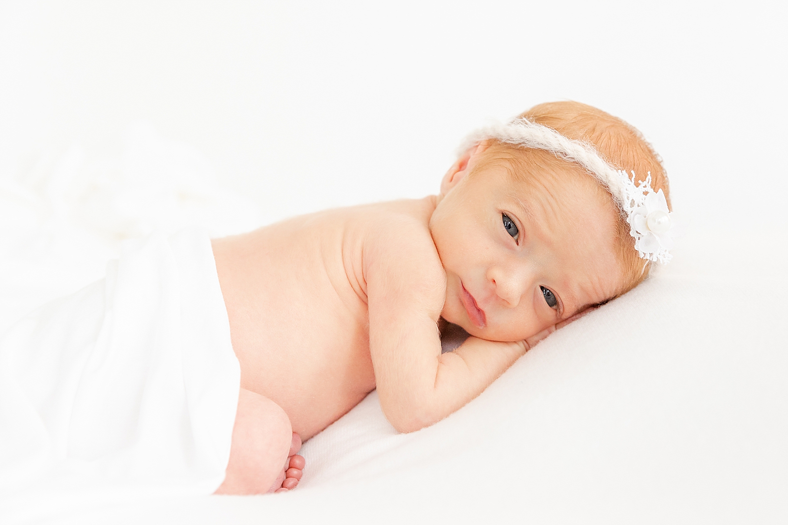 Preemie Newborn Session of posed baby on a white backdrop awake and looking at camera with a white headband on and white blanket draped over her bottom and her toes peeking out