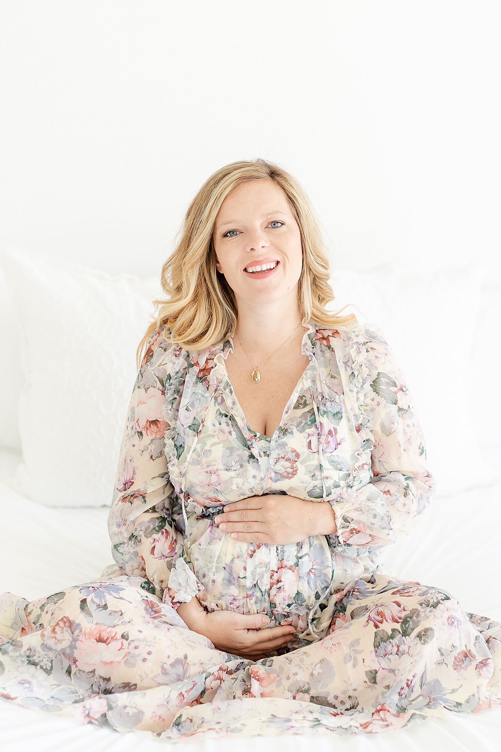 Blonde pregnant mom in floral dress sitting on white bed holding baby bump smiling at camera