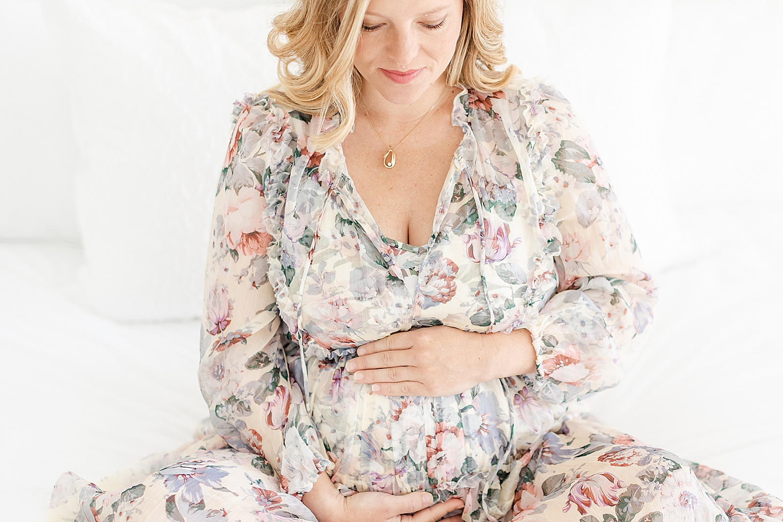 pregnant woman sitting on bed holding belly looking down at her baby wearing floral dress