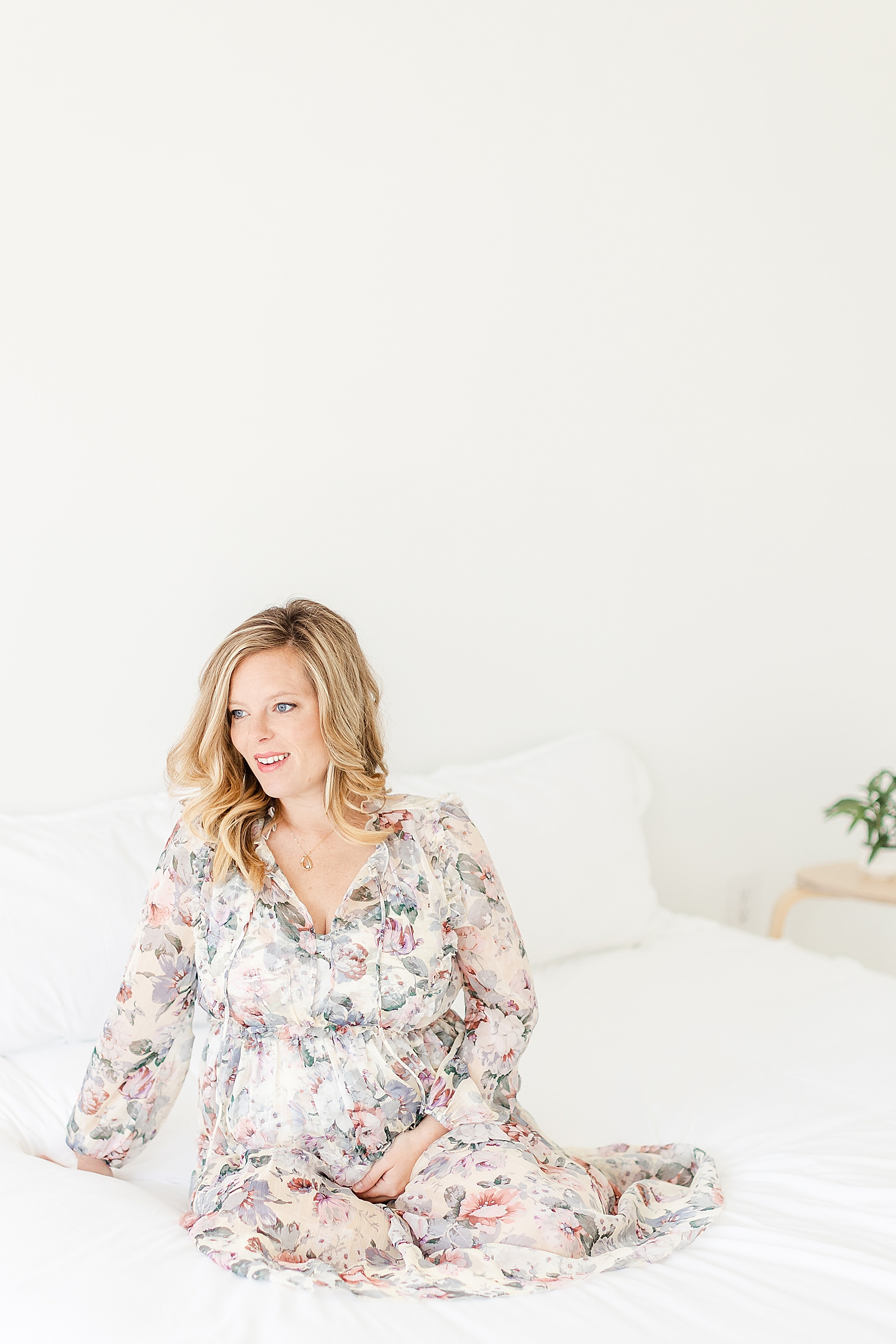 Pregnant mom sitting on bed looking out window holding baby bump wearing floral long sleeve dress for maternity photos