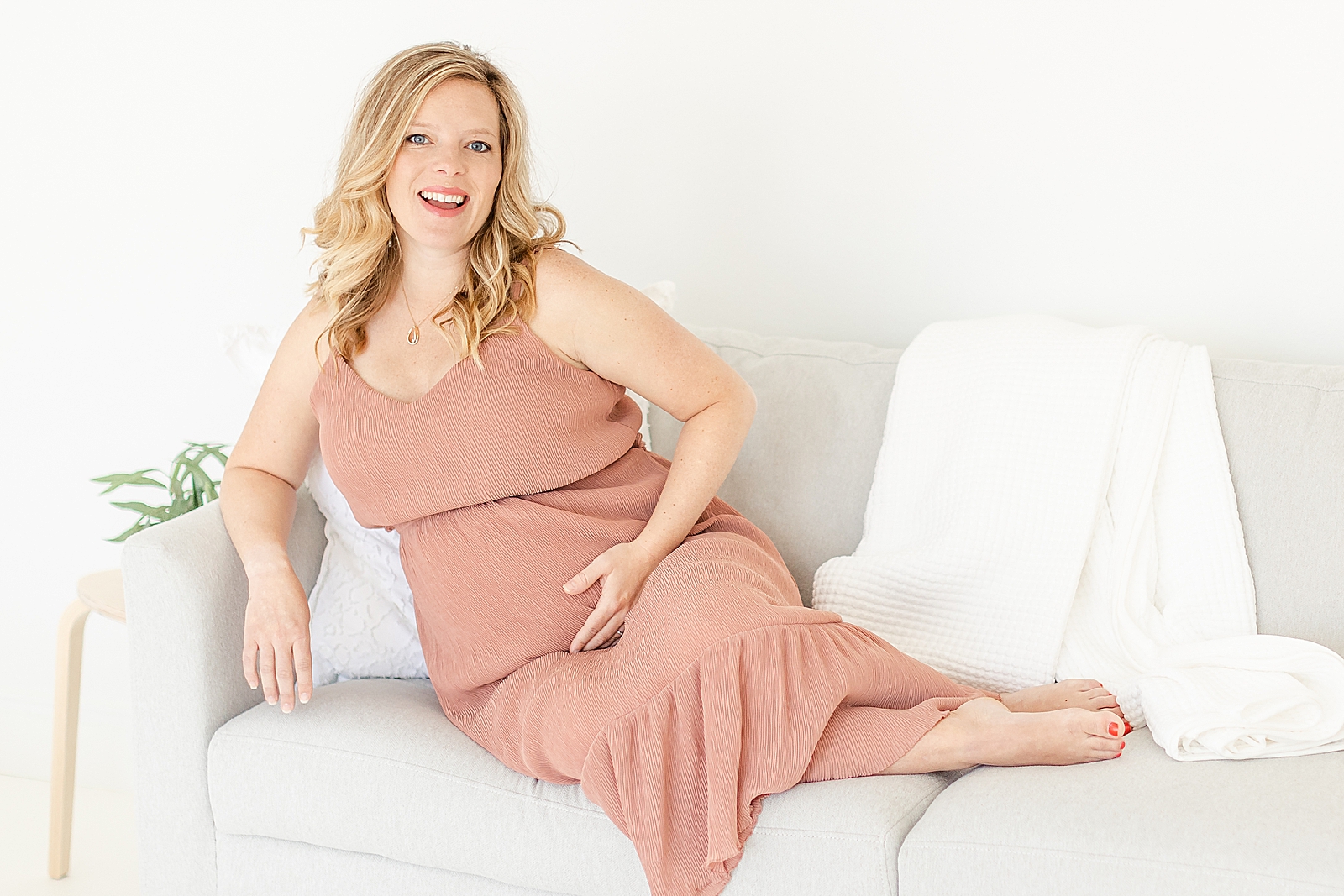 branding maternity photos of mom sitting on grey couch wearing dusty rose dress smiling at camera holding baby bump