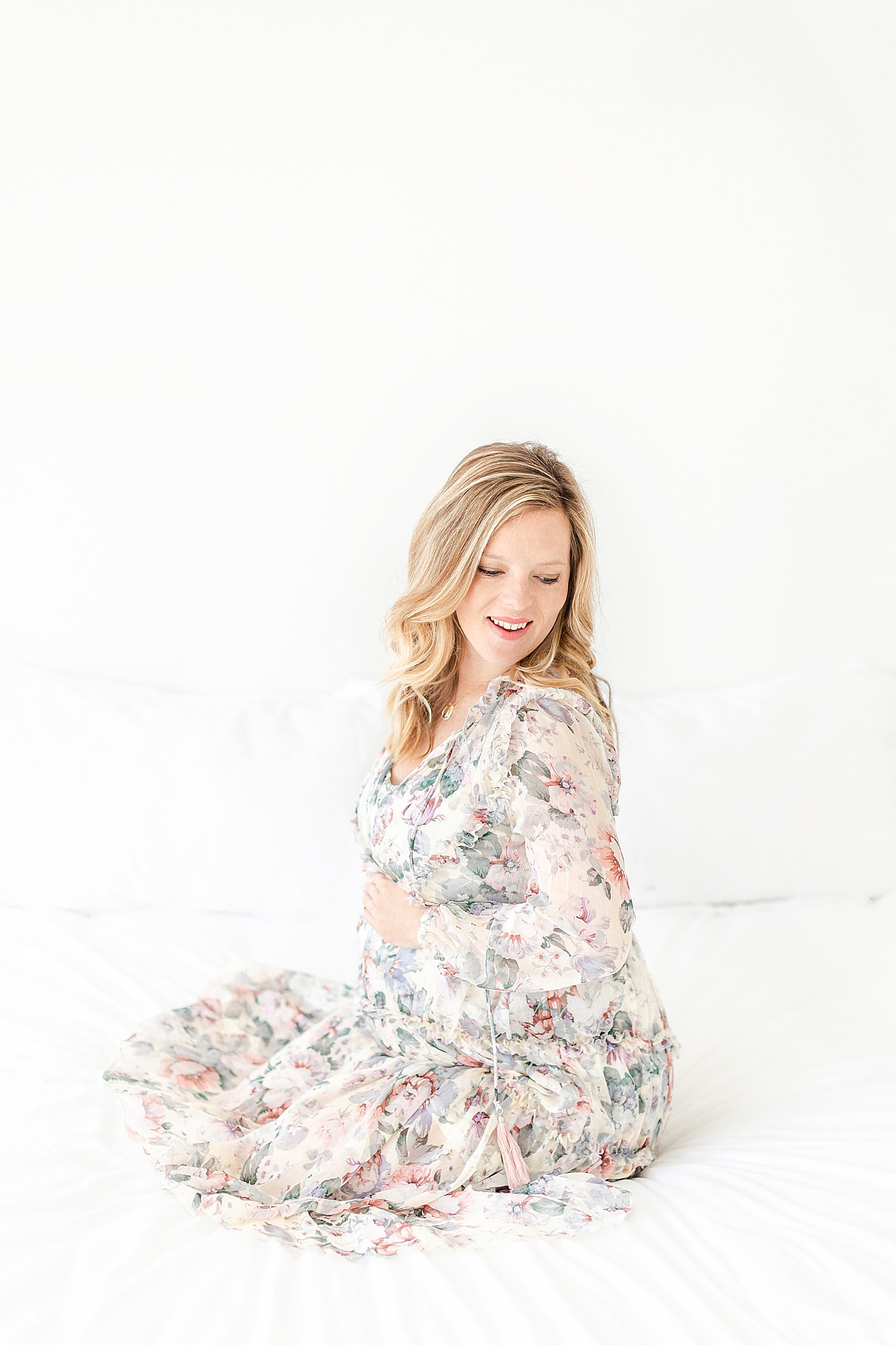 Maternity photo of mom sitting on bed in floral dress holding baby bump and looking down shoulder