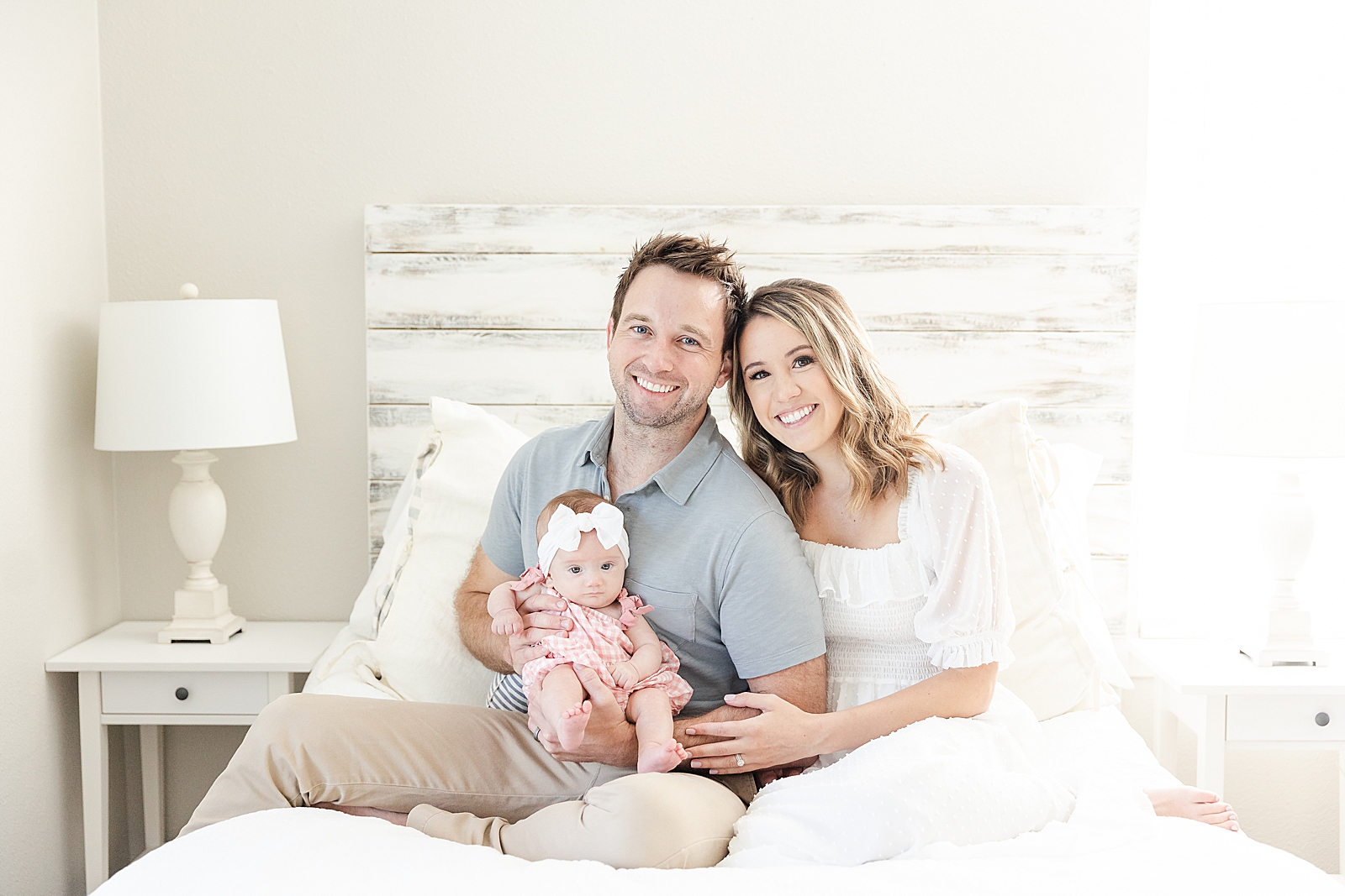 Lifestyle Newborn Photos mom and dad sitting on bet smiling with baby in pink romper and white bow