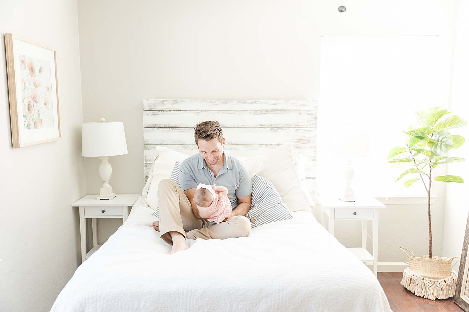 dad sitting in middle of white bed holding out baby and smiling