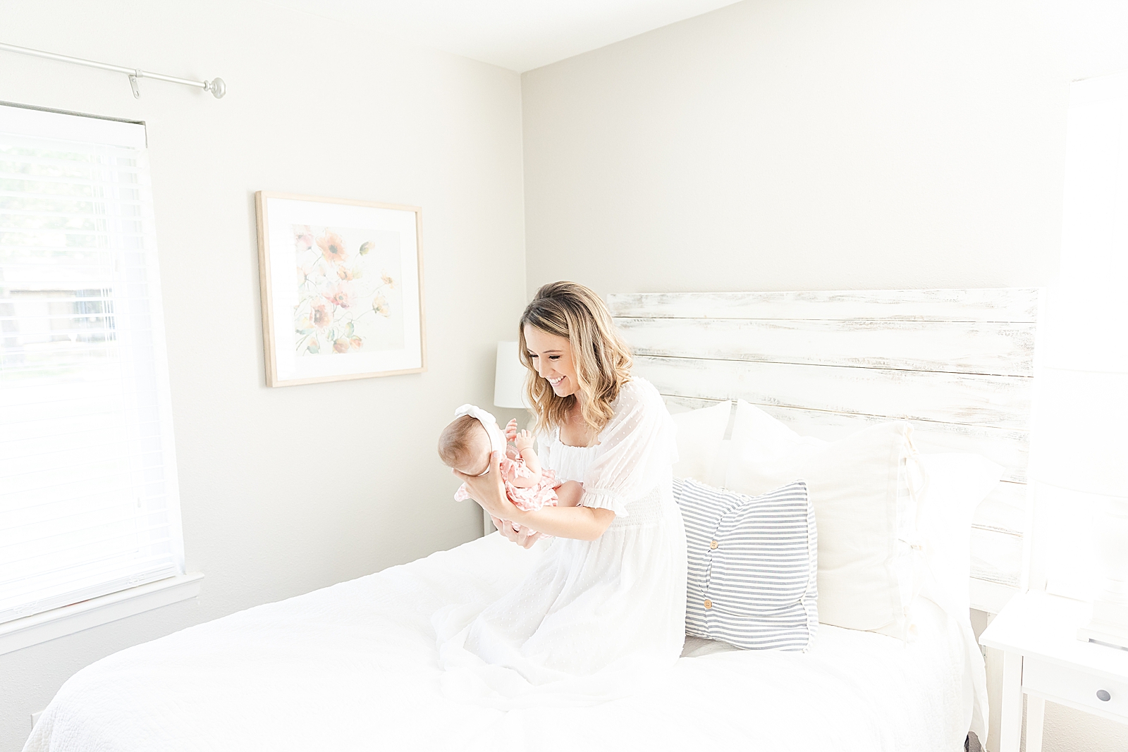 mom on white bed holding and smiling at baby wearing a white dress
