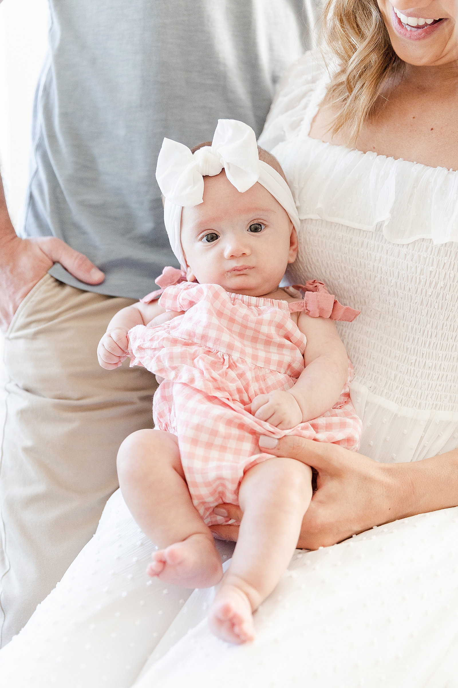 mom holding baby with dad in the background baby with white bow and pink plaid romper sitting and looking at camera