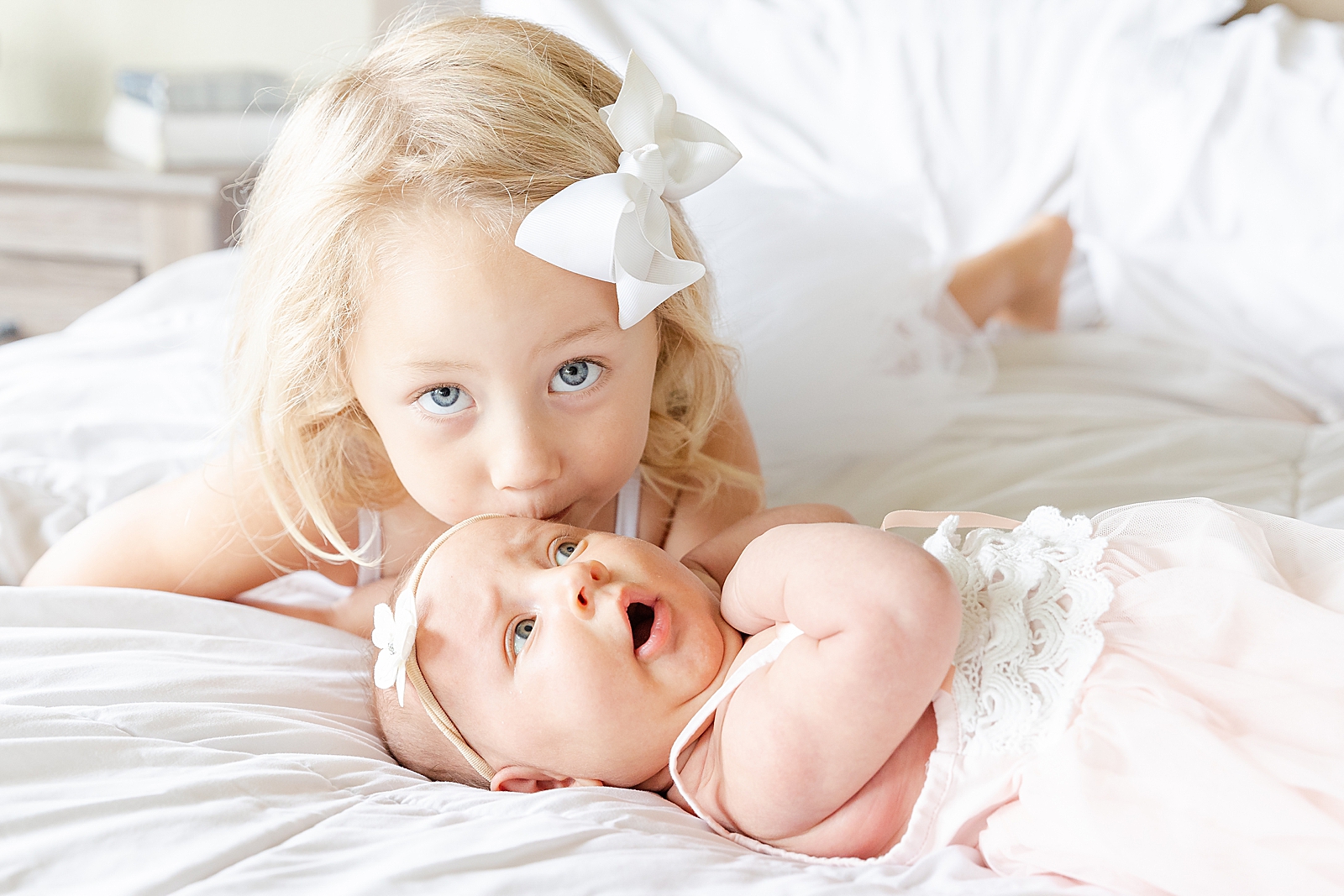 toddler with white bow laying on bed kissing baby sisters head