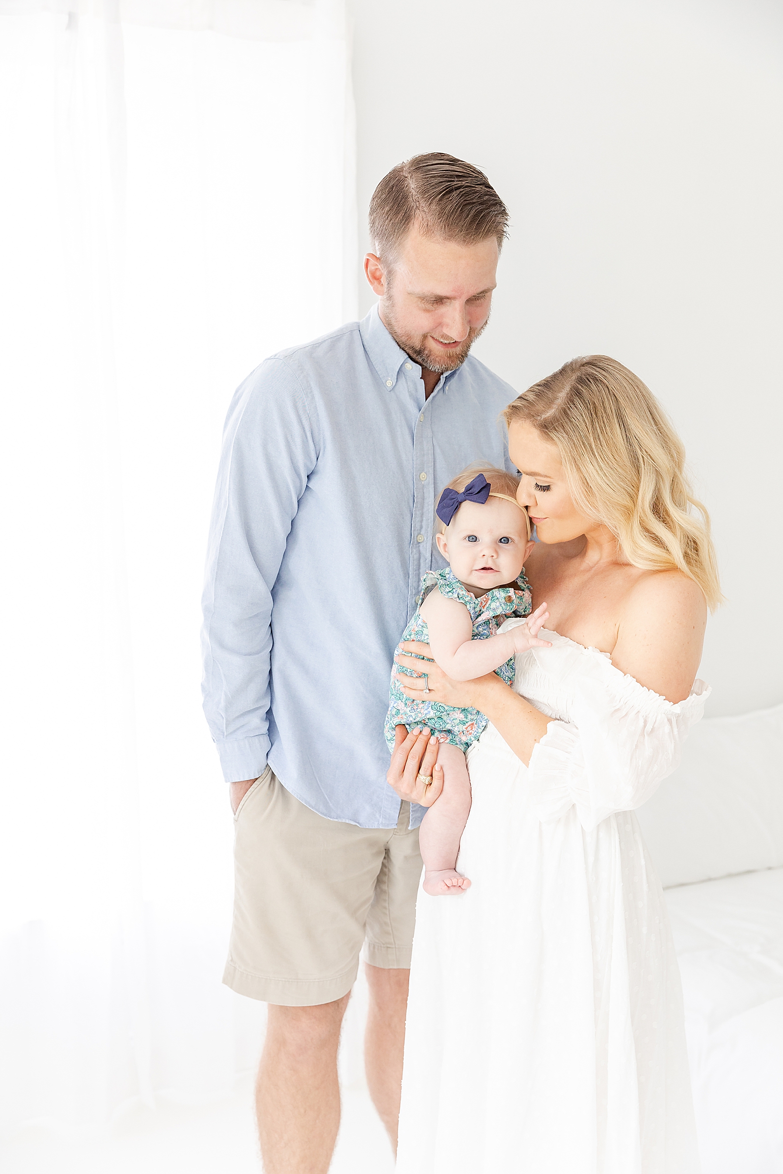 studio family session mom wearing white off the shoulder dress and dad wearing flue shirt and kaki short standing looking at baby smiling