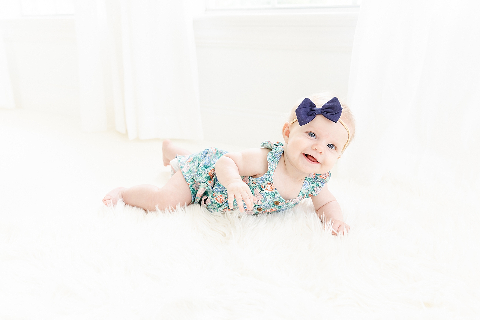 baby girl in floral romper from old navy with a blue bow on tummy on white fur rug smiling up at camera
