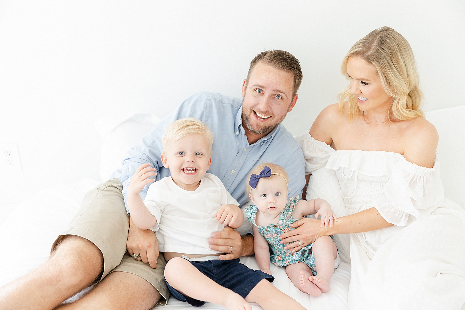 Studio family session on white bed dad baby and toddler smiling at camera while mom smiles at family