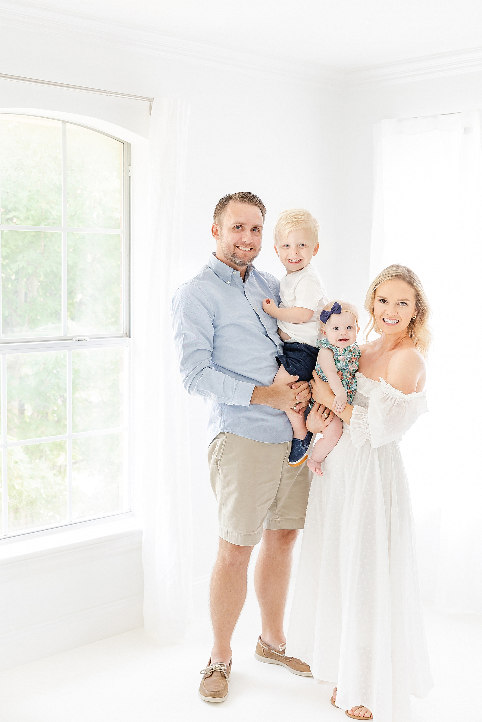 family photo standing nest to a window in a white studio mom wearing white dress everyone smiling at camera