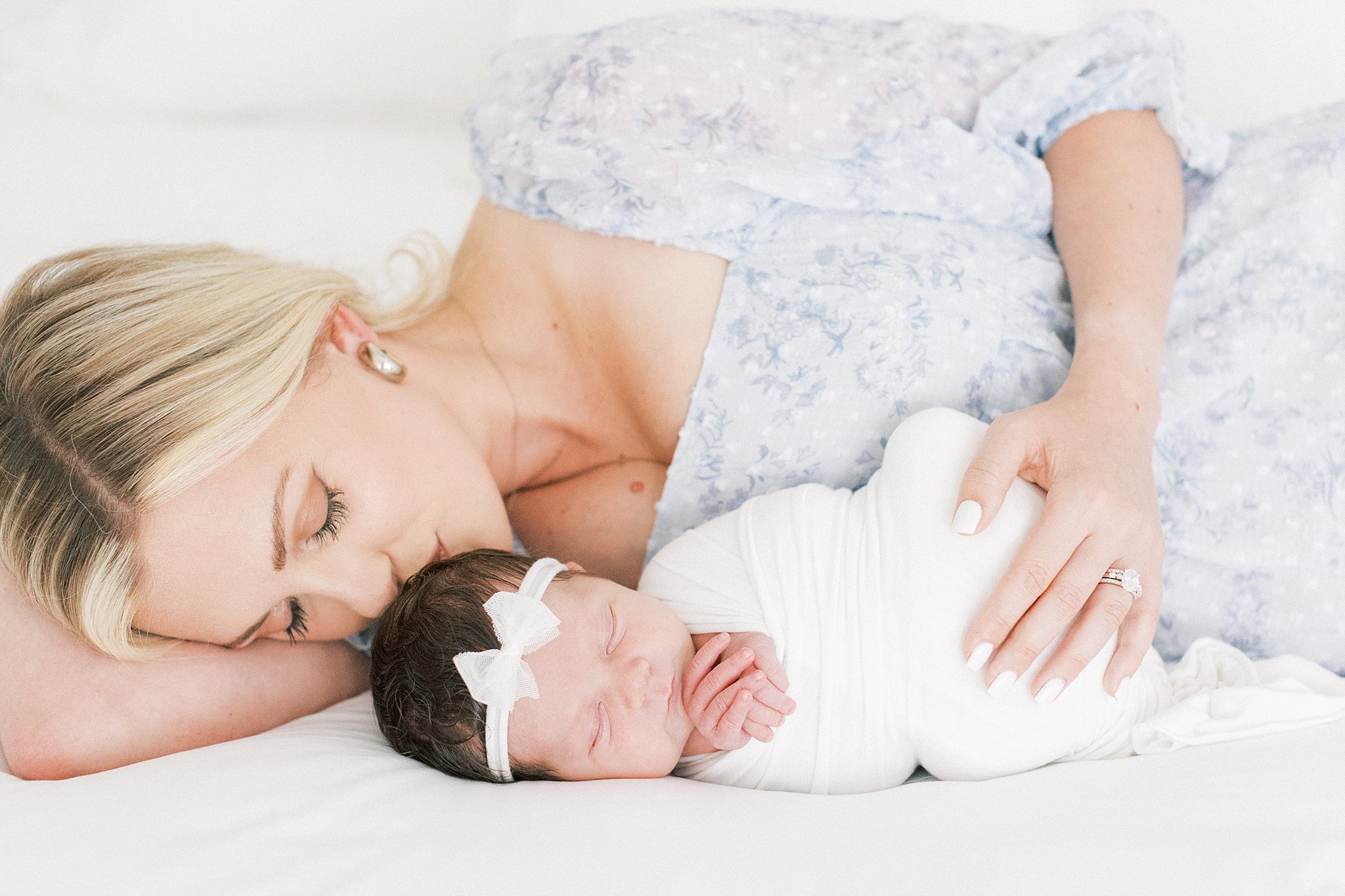 A mother cuddles alongside her sleeping newborn daughter wearing a white swaddle and white bow headband Dallas baby boutiques
