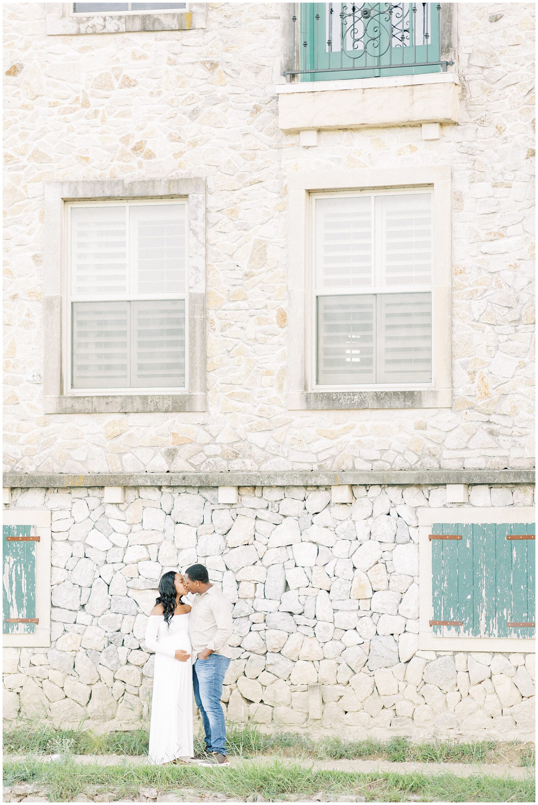 Parents to be stand kissing on a sidewalk in front of a historical stone building with green shutters