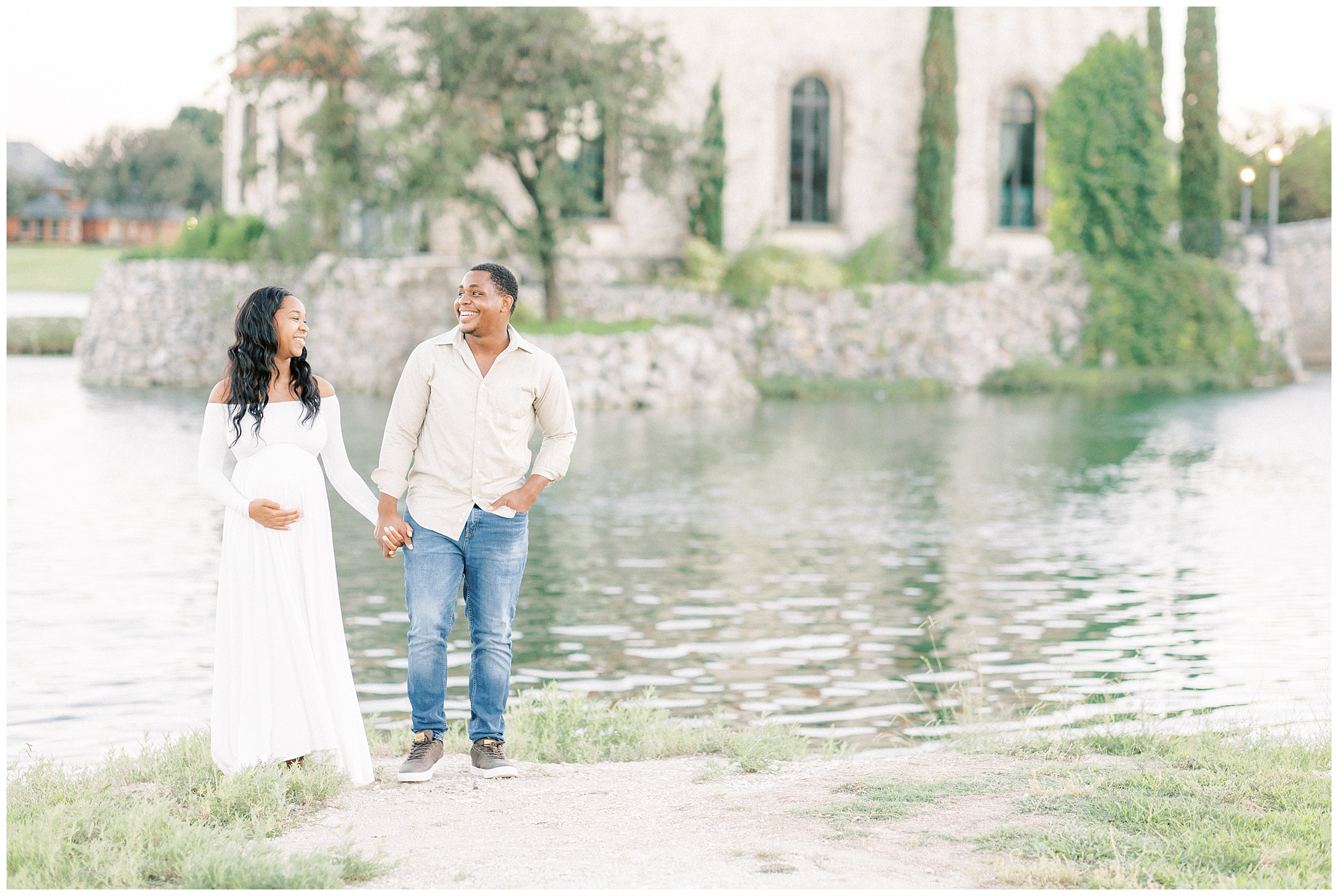 A mother to be walks along a lake with a stone castle in the background while holding hands with her partner wearing jeans and a tan button down Dallas Midwives