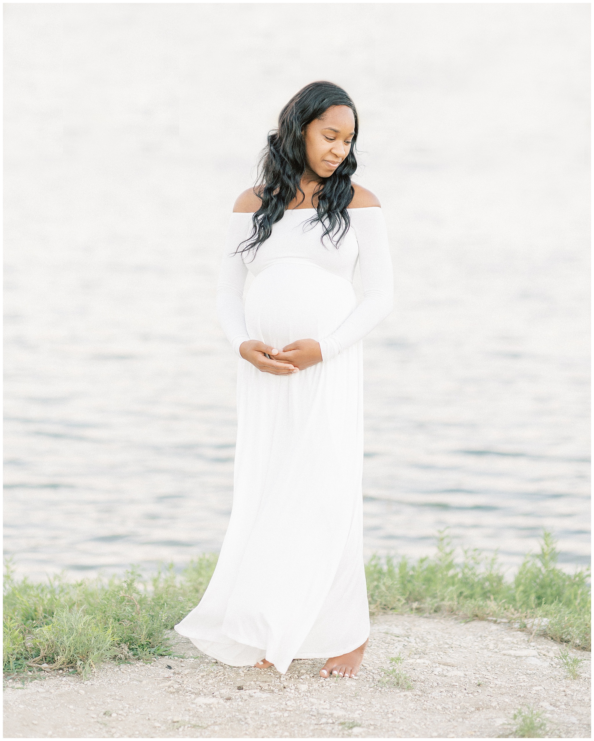 A mother to be in a white maternity gown stands barefoot on the edge of a lake holding her bump Dallas Midwives