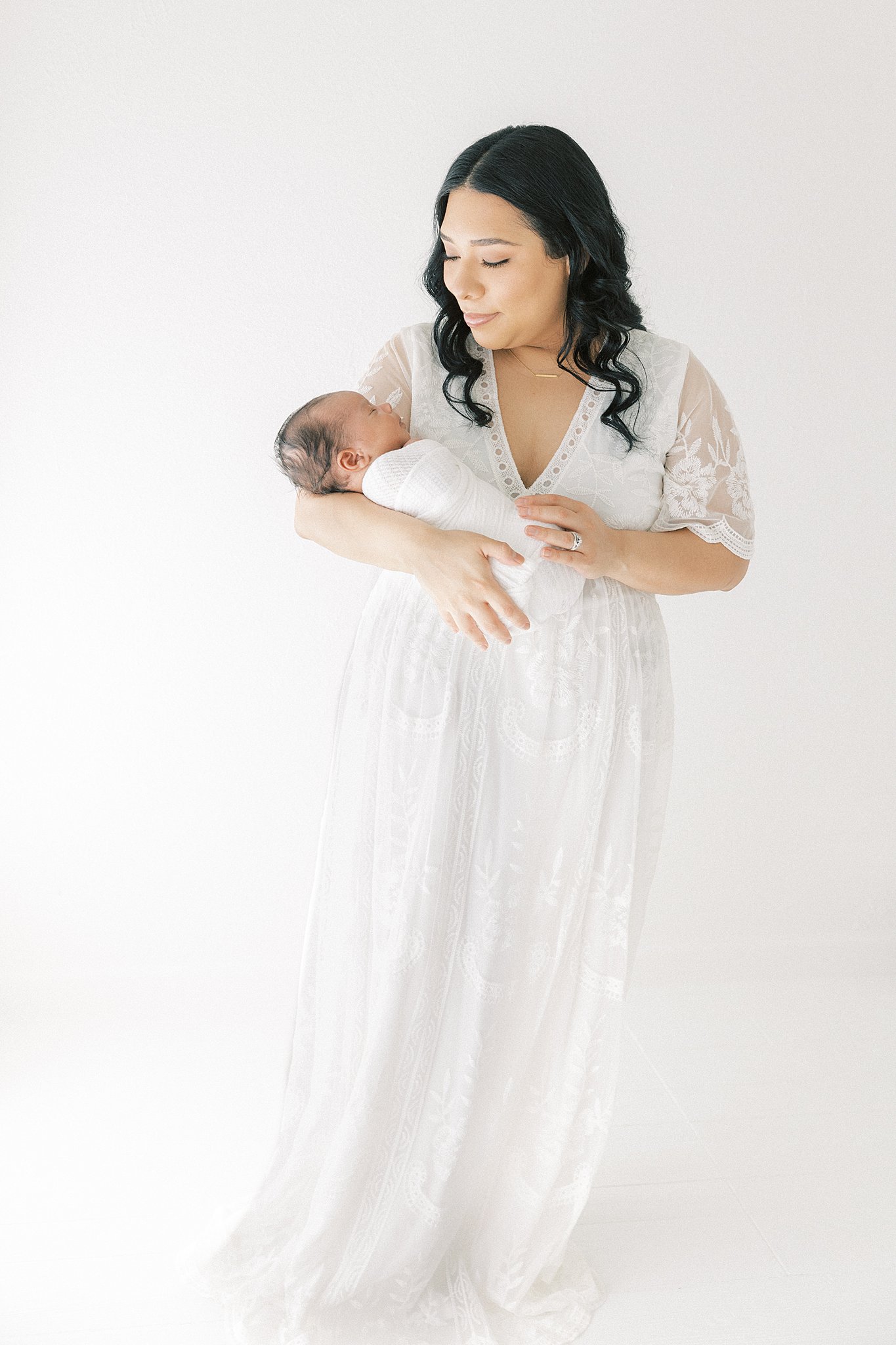 A mother in a white lace dress stands in a studio holding her newborn baby boy