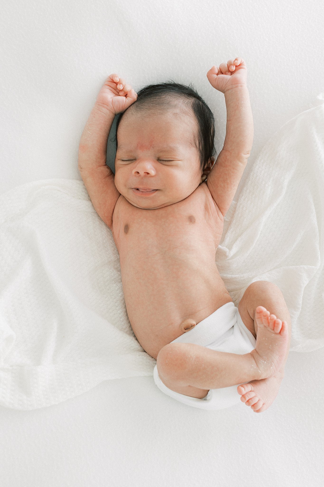 A newborn baby stretches it's arms way above it's head on a white bed baby bliss Dallas