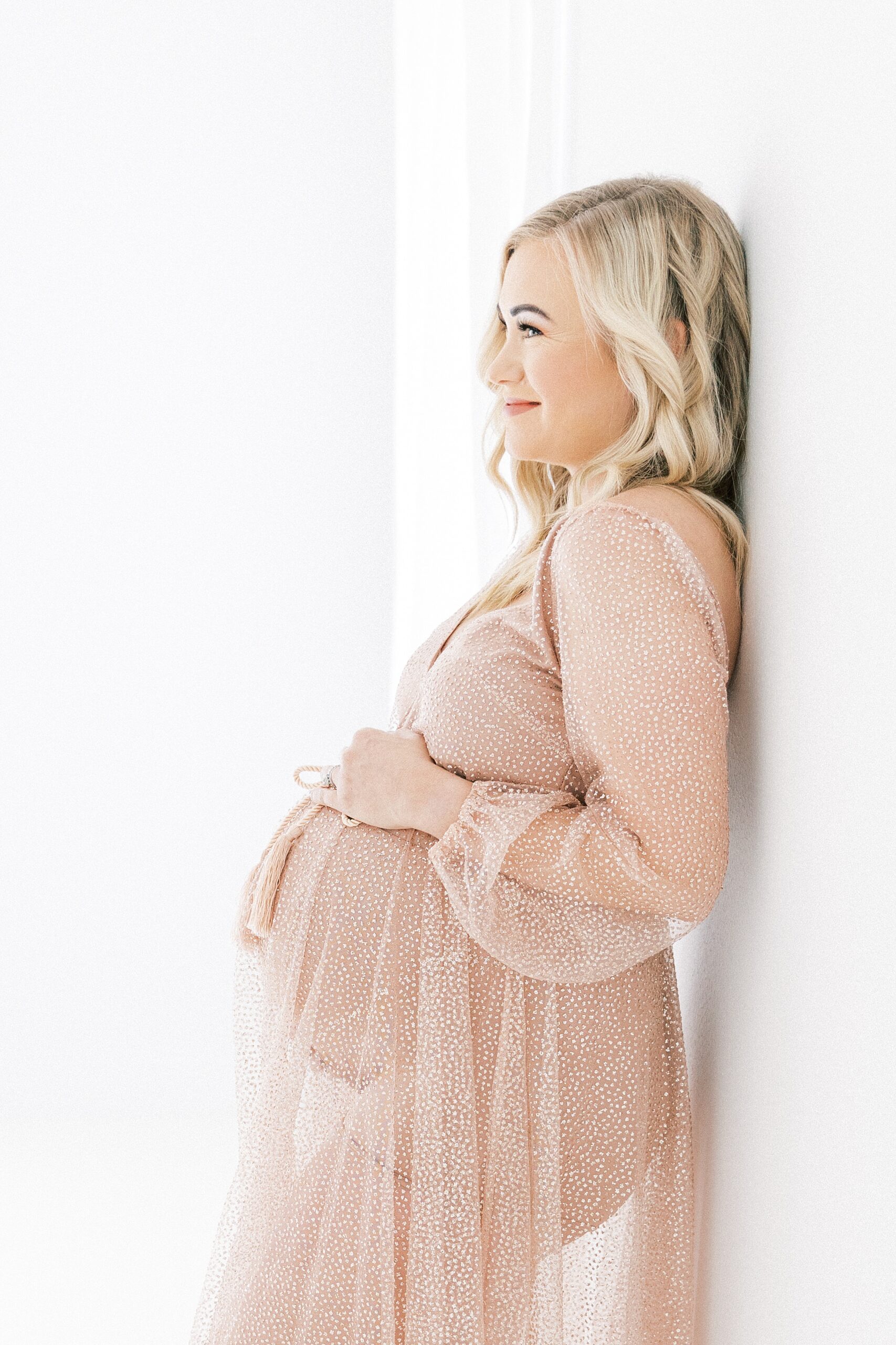 A mom to be in a beige coverup leans against a wall in a studio dallas birth center