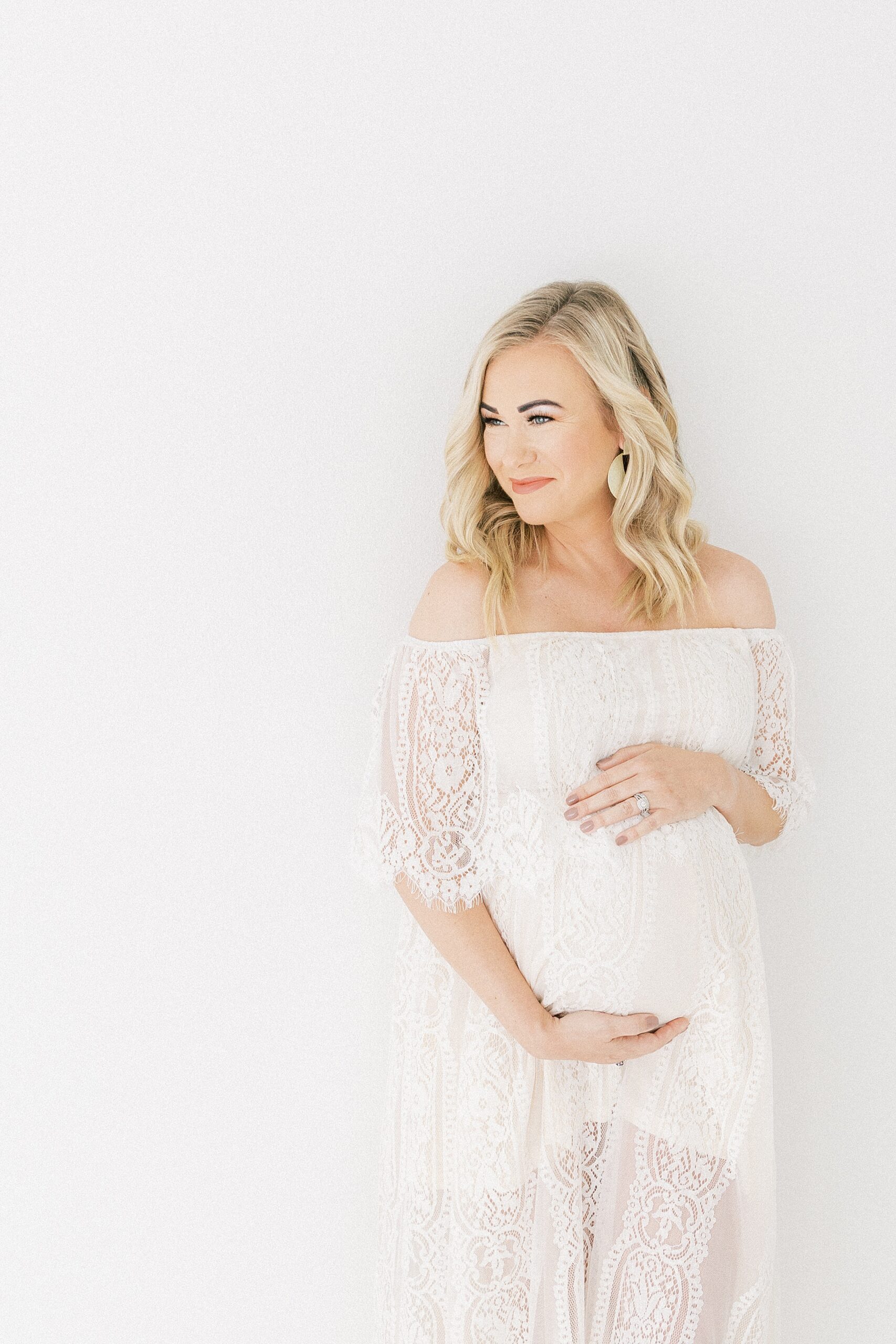 A pregnant woman with blonde hair stands in a studio in a white lace maternity dress dallas birth center
