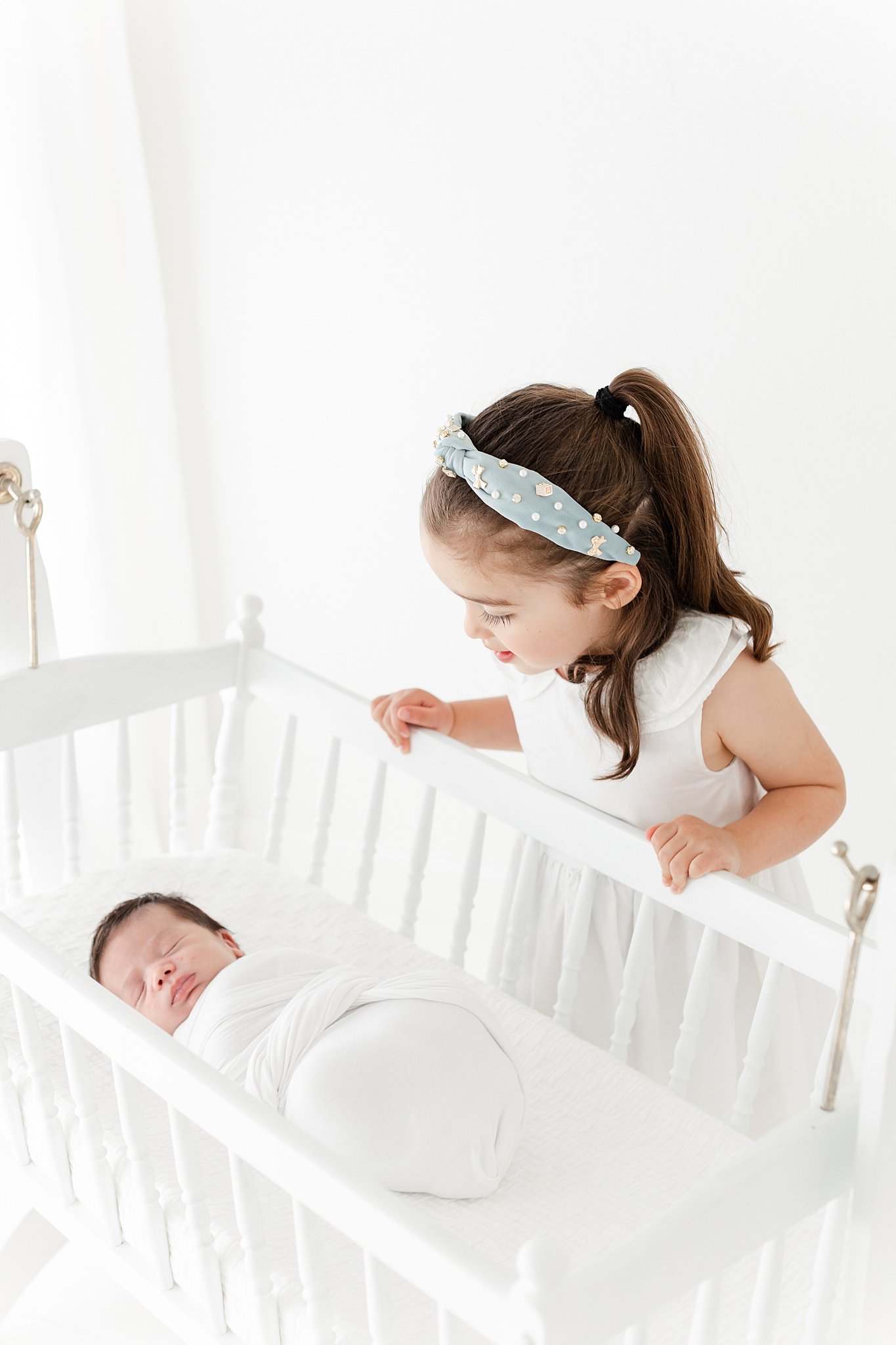 A young girl leans over the crib to see her sleeping newborn sibling