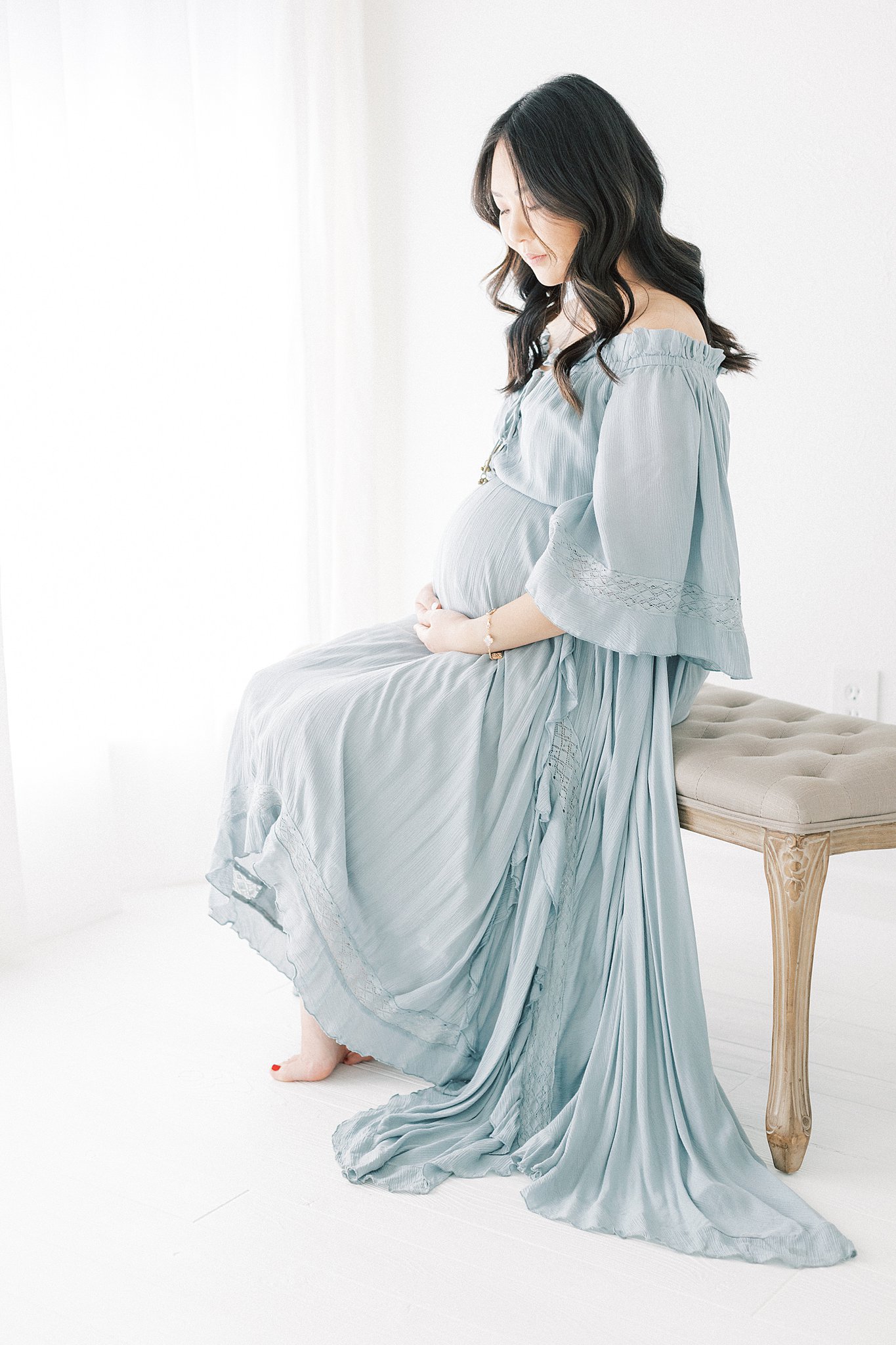 A mother to be sits on a bench in a studio by a window in a blue maternity gown looking down at her bump