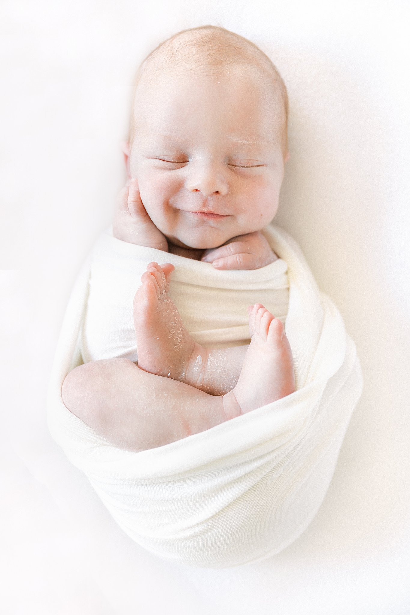 A newborn baby smiles in its sleep while laying in a white swaddle Jojo Mommy