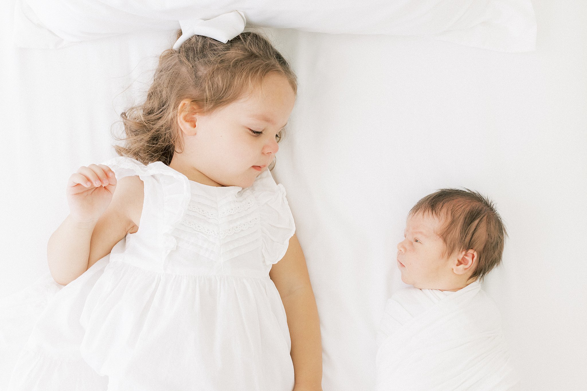 A young girl in a white dress lays on a bed with her newborn baby sibling wrapped in a white swaddle layette dallas